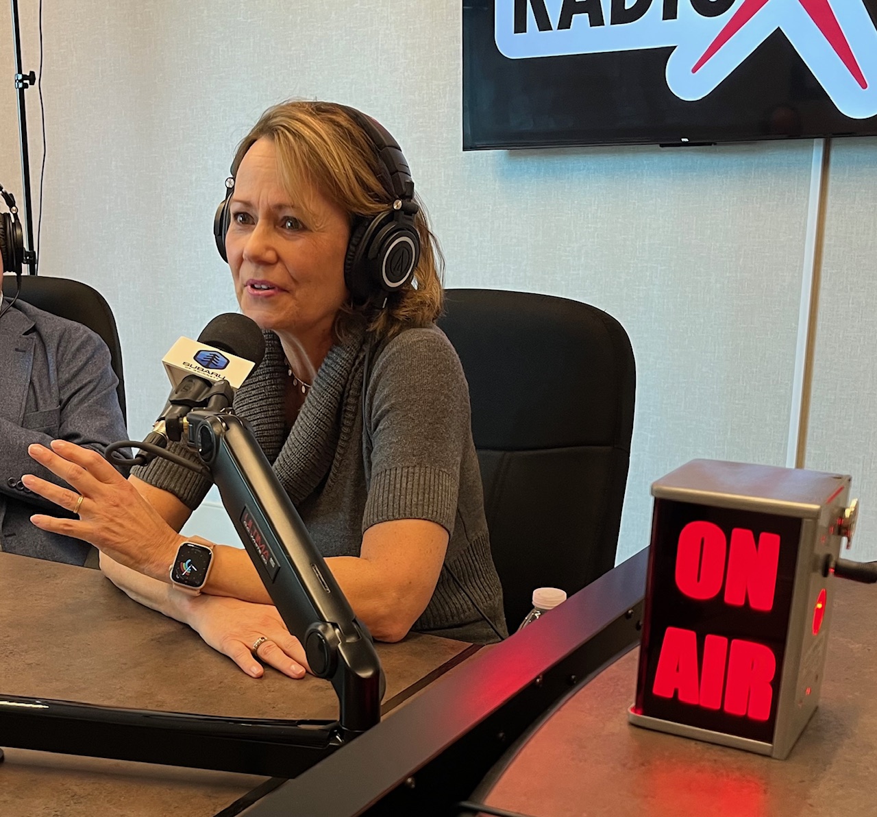 Woman with headphones speaking into microphone during radio broadcast, with ON AIR sign visible.