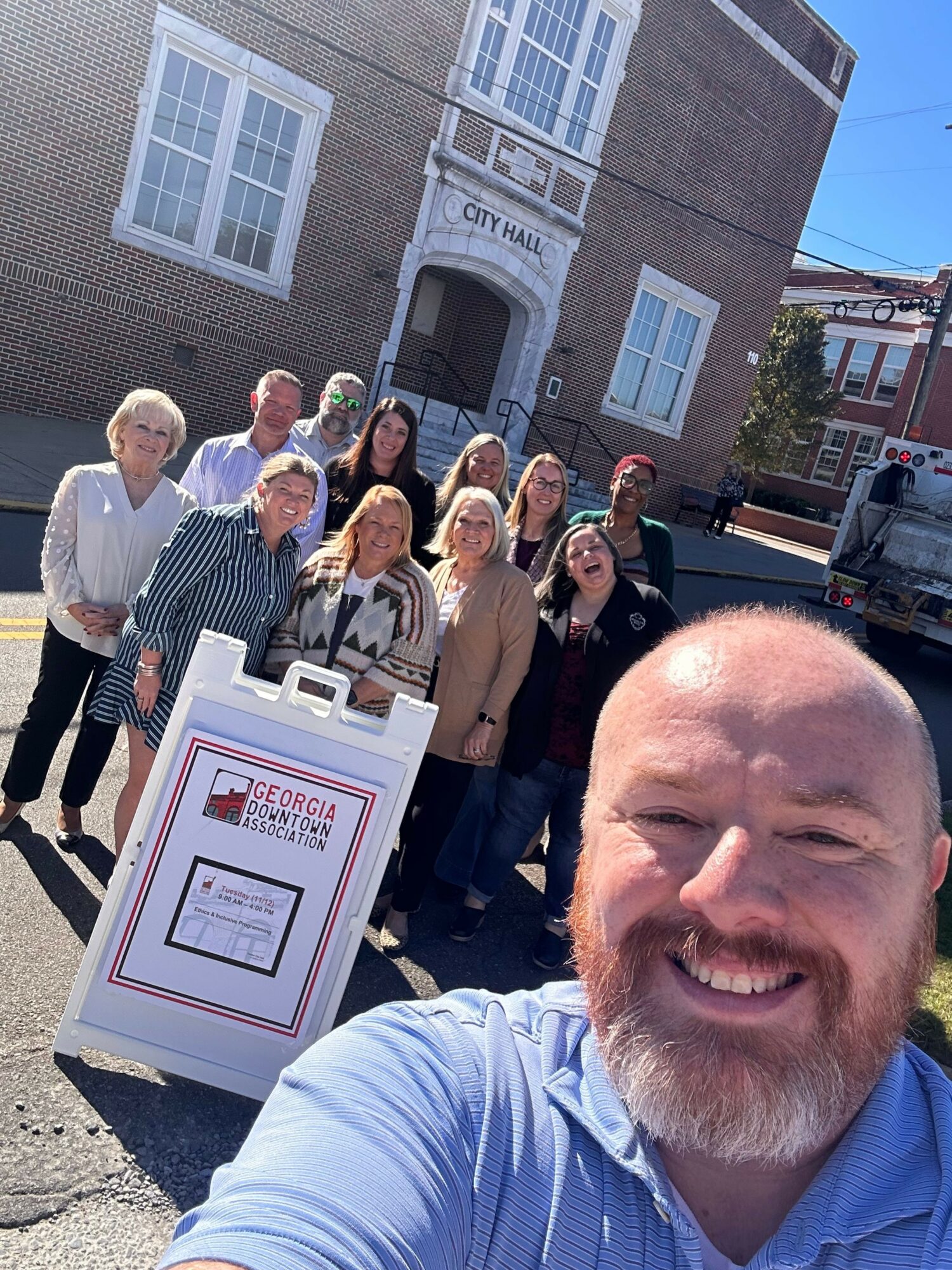 Group of nine people standing outside a brick building with a sign reading 'City Hall'. A man in the foreground takes a selfie.