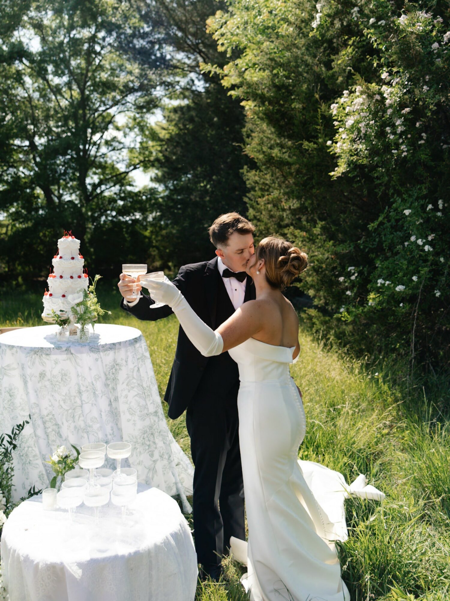 Bride and groom dancing outdoors with wedding cake and tables nearby, surrounded by trees and greenery.