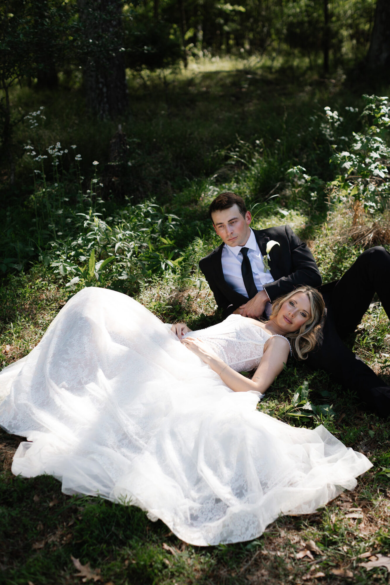 A bride and groom lying on grass in a wooded area, bride in a white gown, groom in a suit.