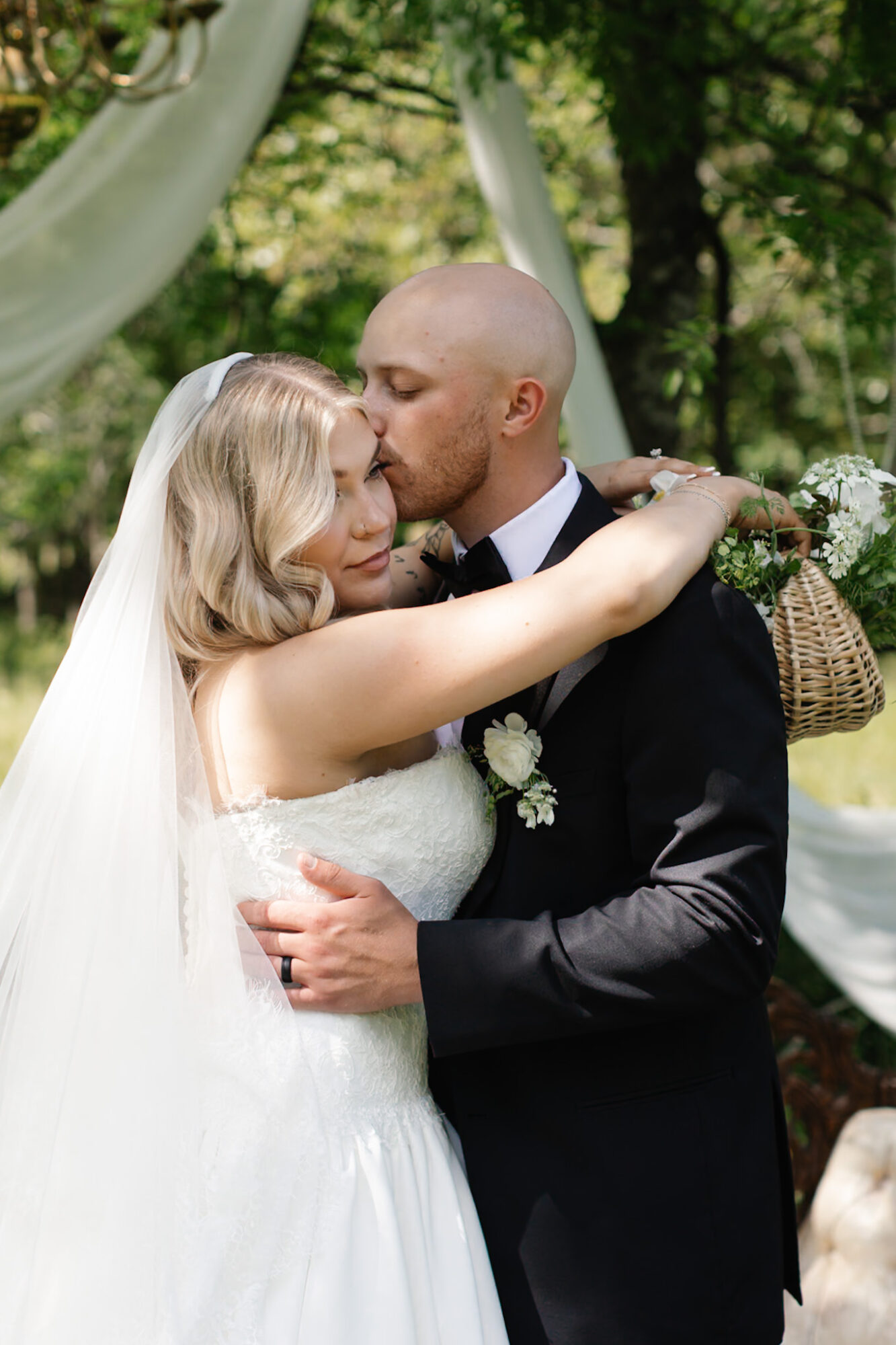 Bride and groom embrace outdoors, bride with arms around groom's neck, groom holding bride, trees and drapery in background.