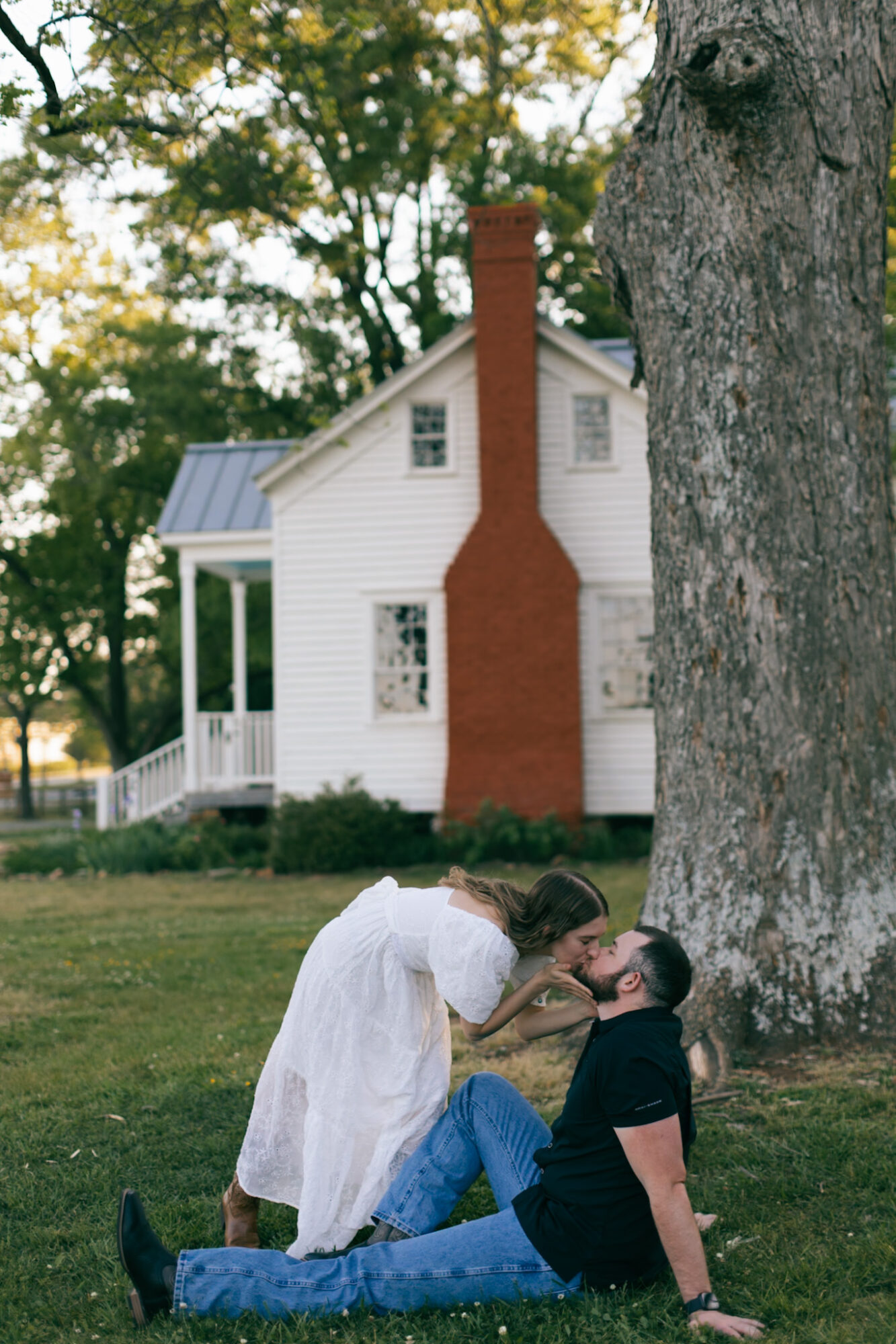 Two women sitting on grass near a large tree, one leaning in to kiss the other, house in background.