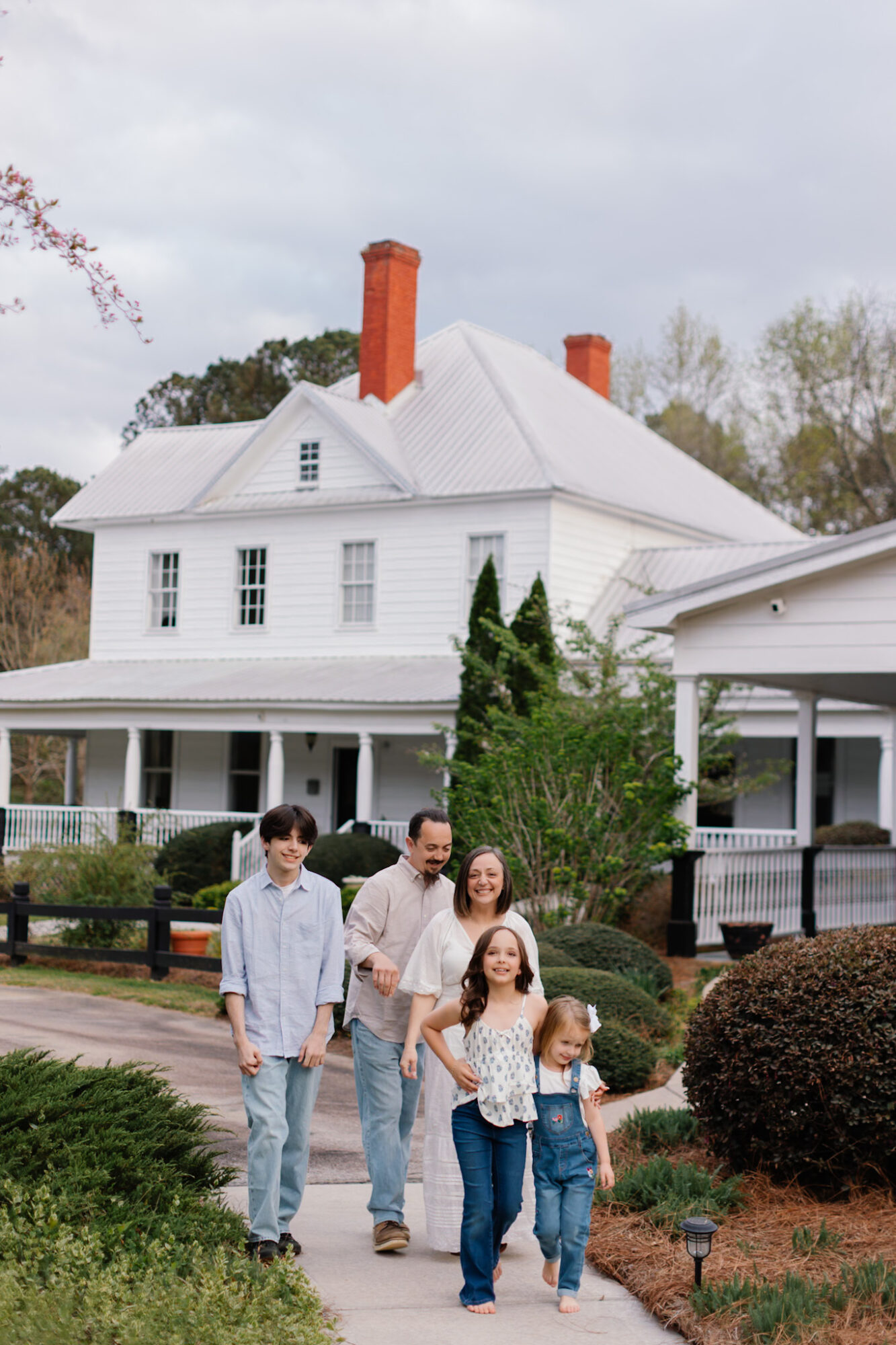 Family of five walking on a sidewalk in front of a large white house with a porch and chimney, trees and bushes around.