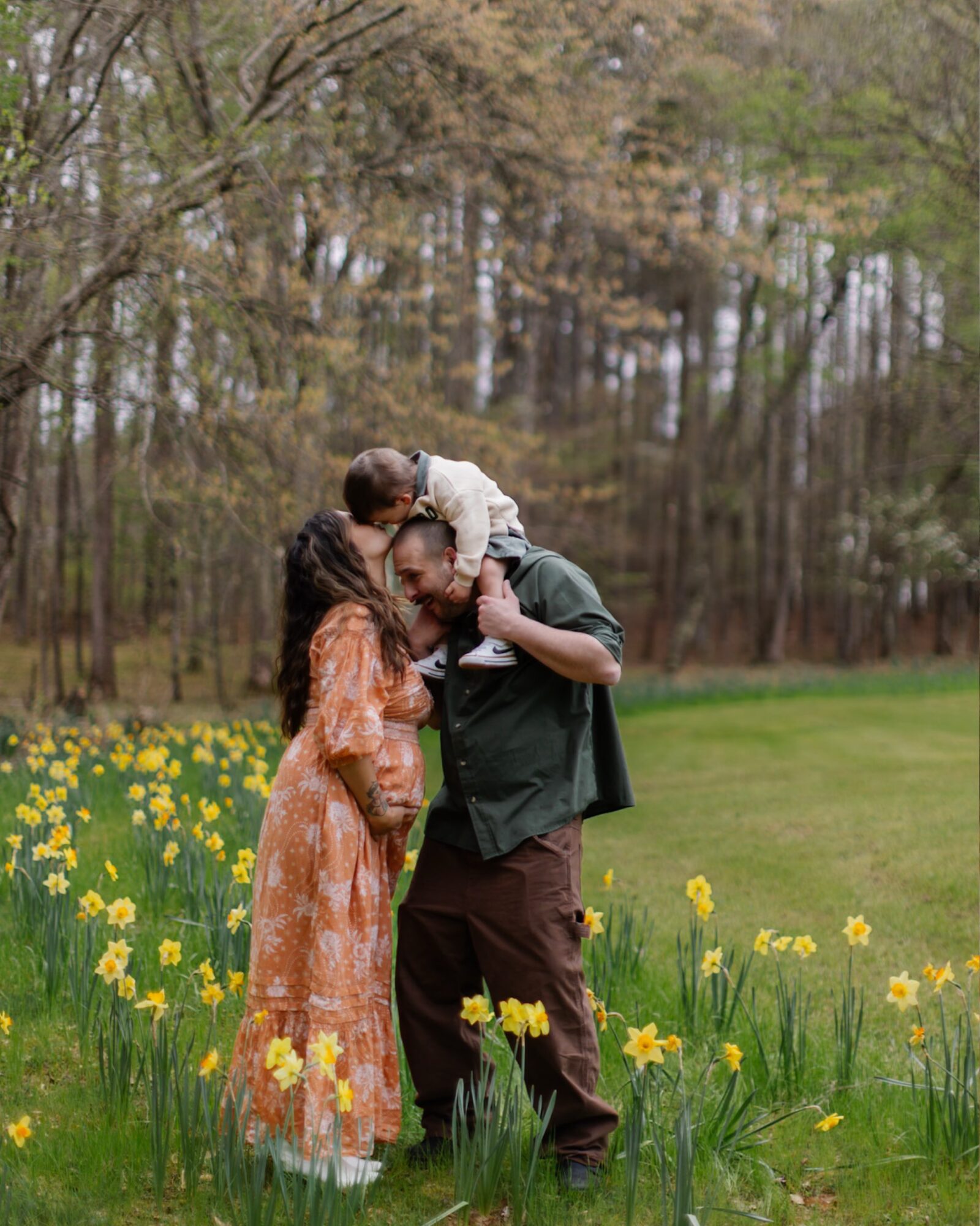 Family outdoors in a field of yellow flowers with trees in the background, a man carrying a child on his shoulders, woman beside him.