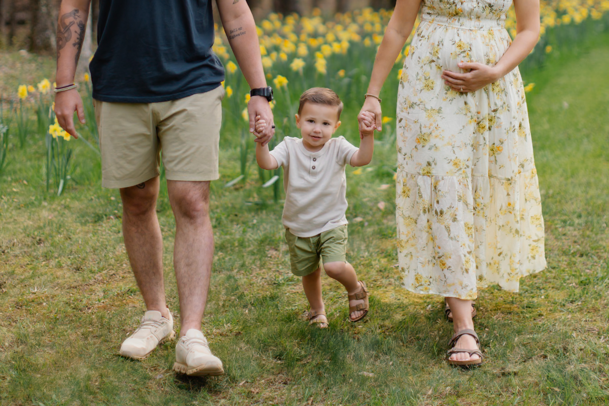 Child holding hands with two adults outdoors on grass with yellow flowers in background.