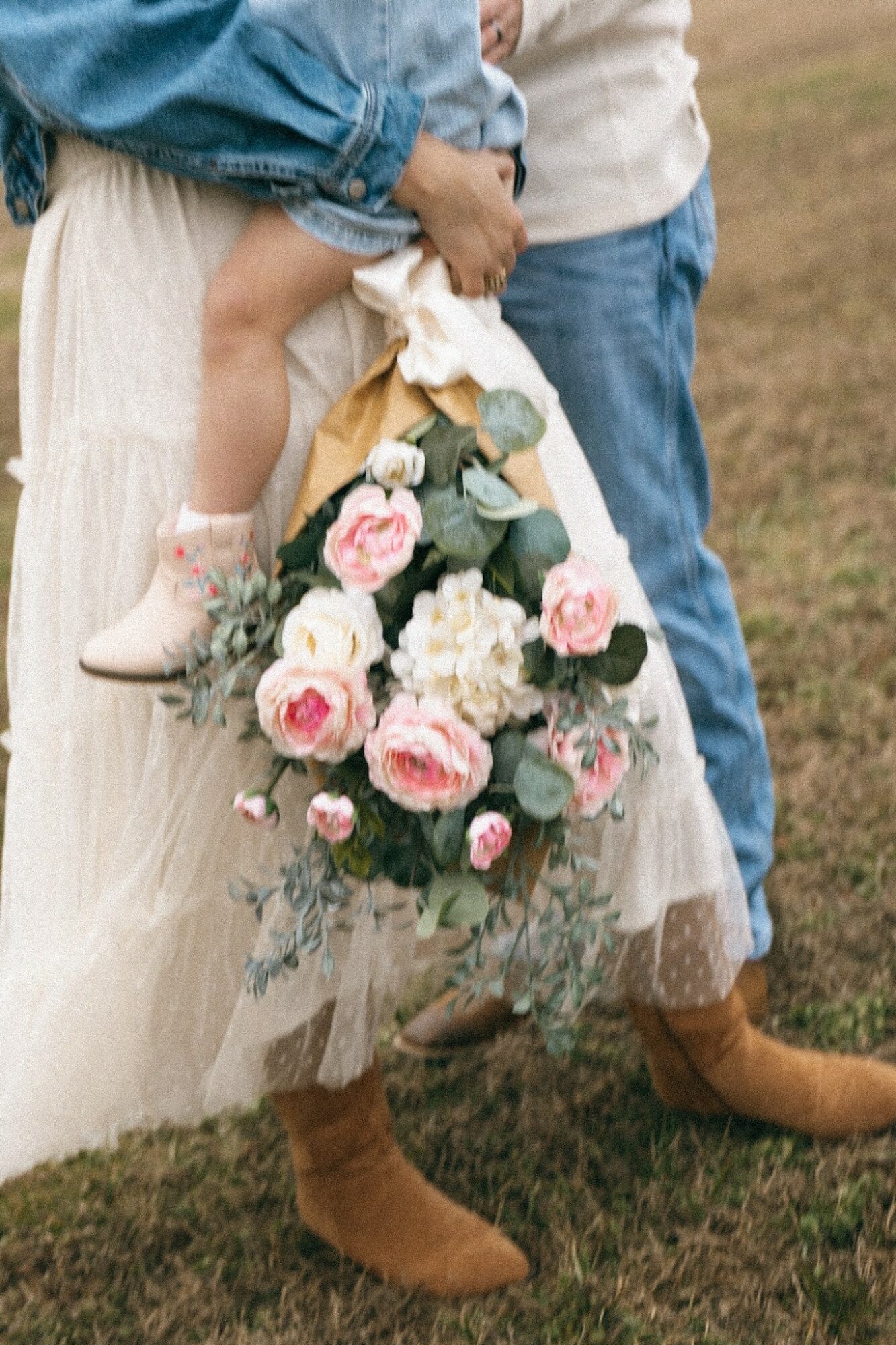 Person holding a child with a bouquet of pink and white roses, standing outdoors on grass.