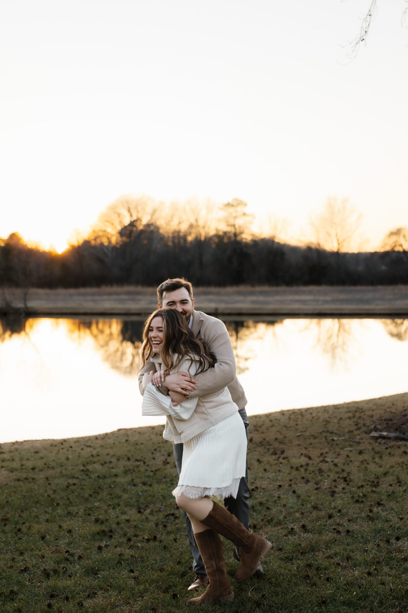 A couple stands near a lake at sunset, embracing and smiling, with trees and sky in the background.
