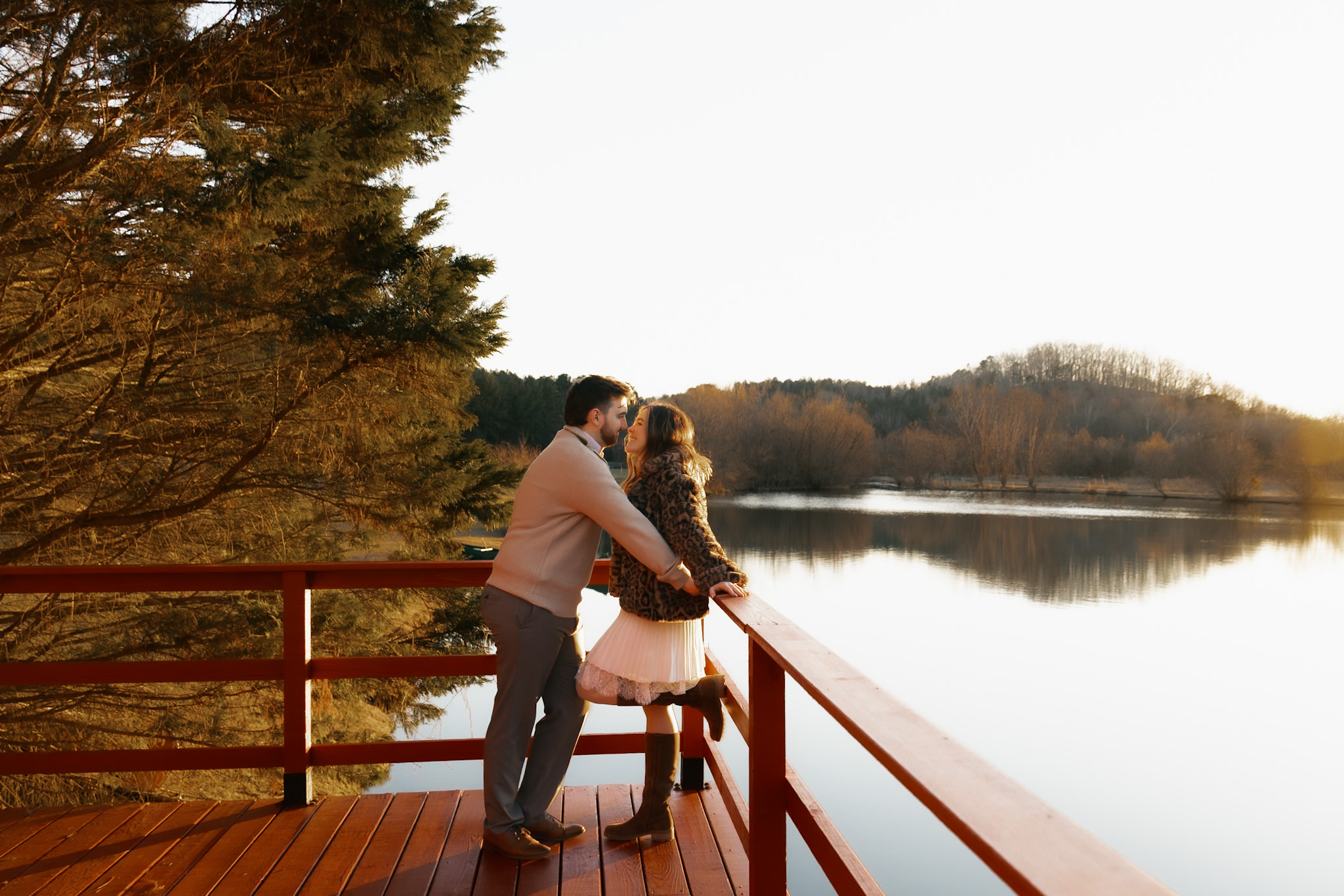 Two people stand on a wooden bridge by a lake, embracing and looking at each other during sunset.