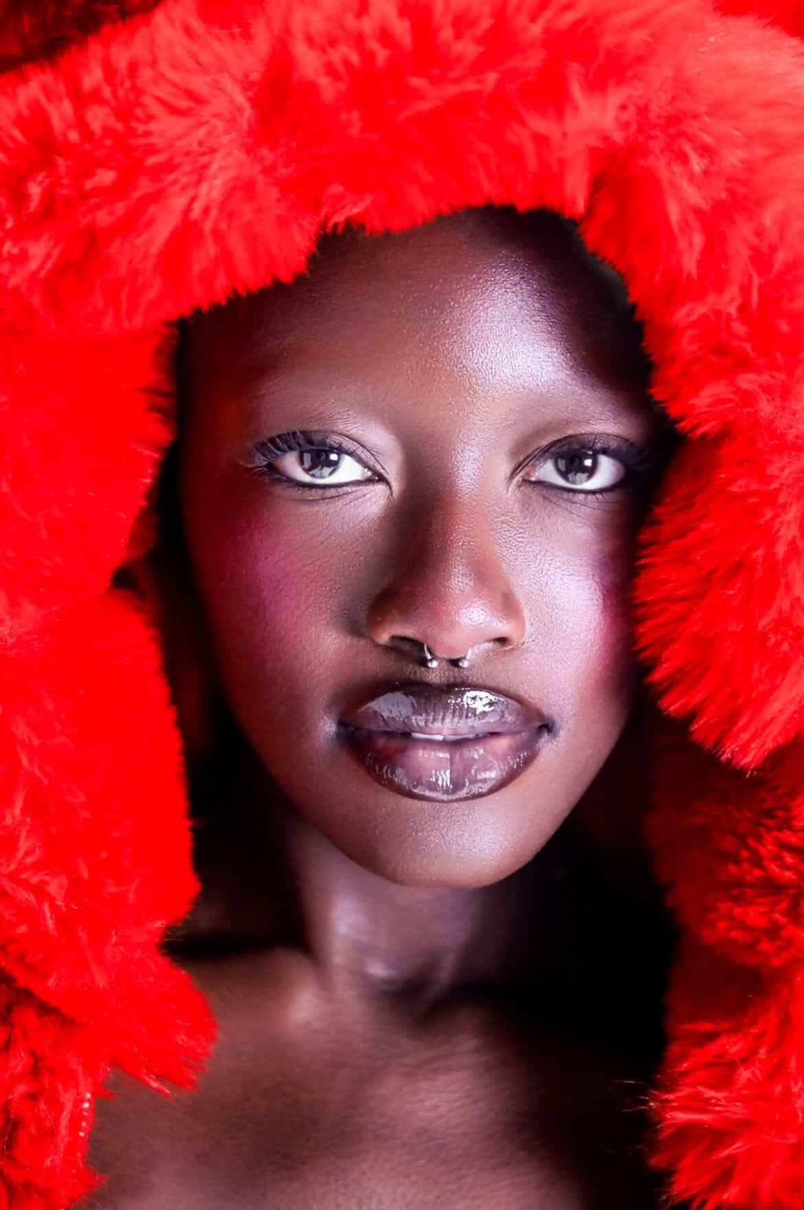 Close-up of a person with dark skin, wearing a red furry hood, with makeup and a septum piercing.