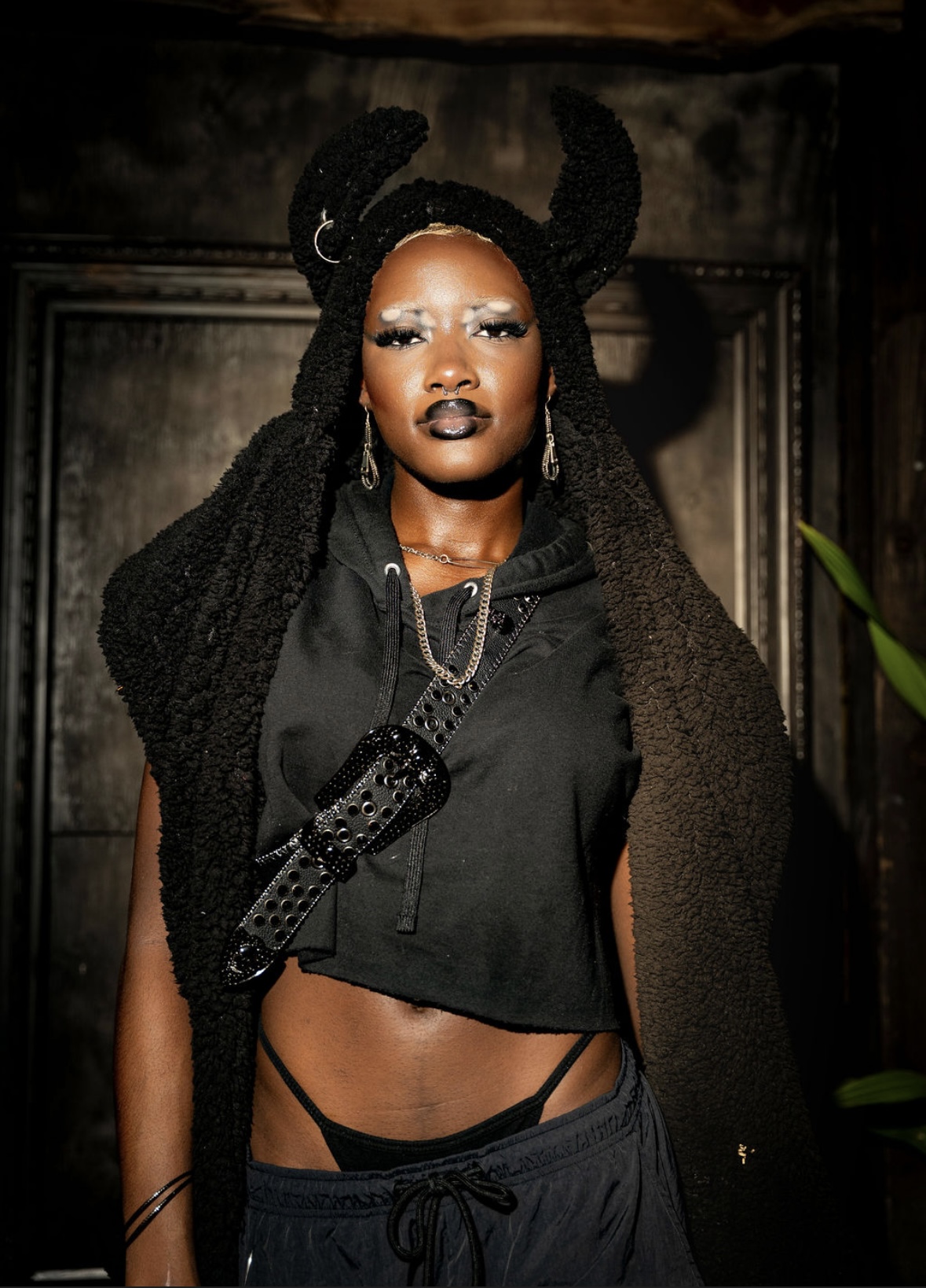 Woman with braided hair and dark makeup wearing a black cropped hoodie and accessories, standing indoors.