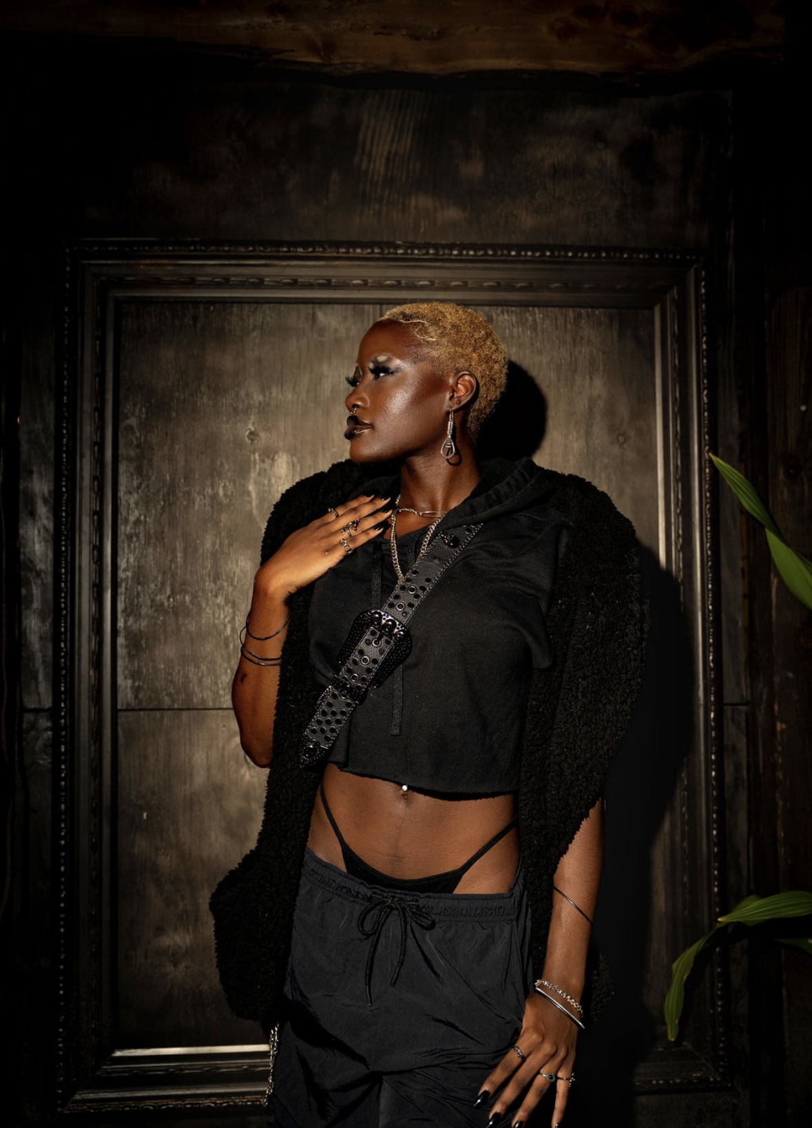 Woman with short blonde hair, wearing black crop top and pants, standing against dark wall, looking to the side.