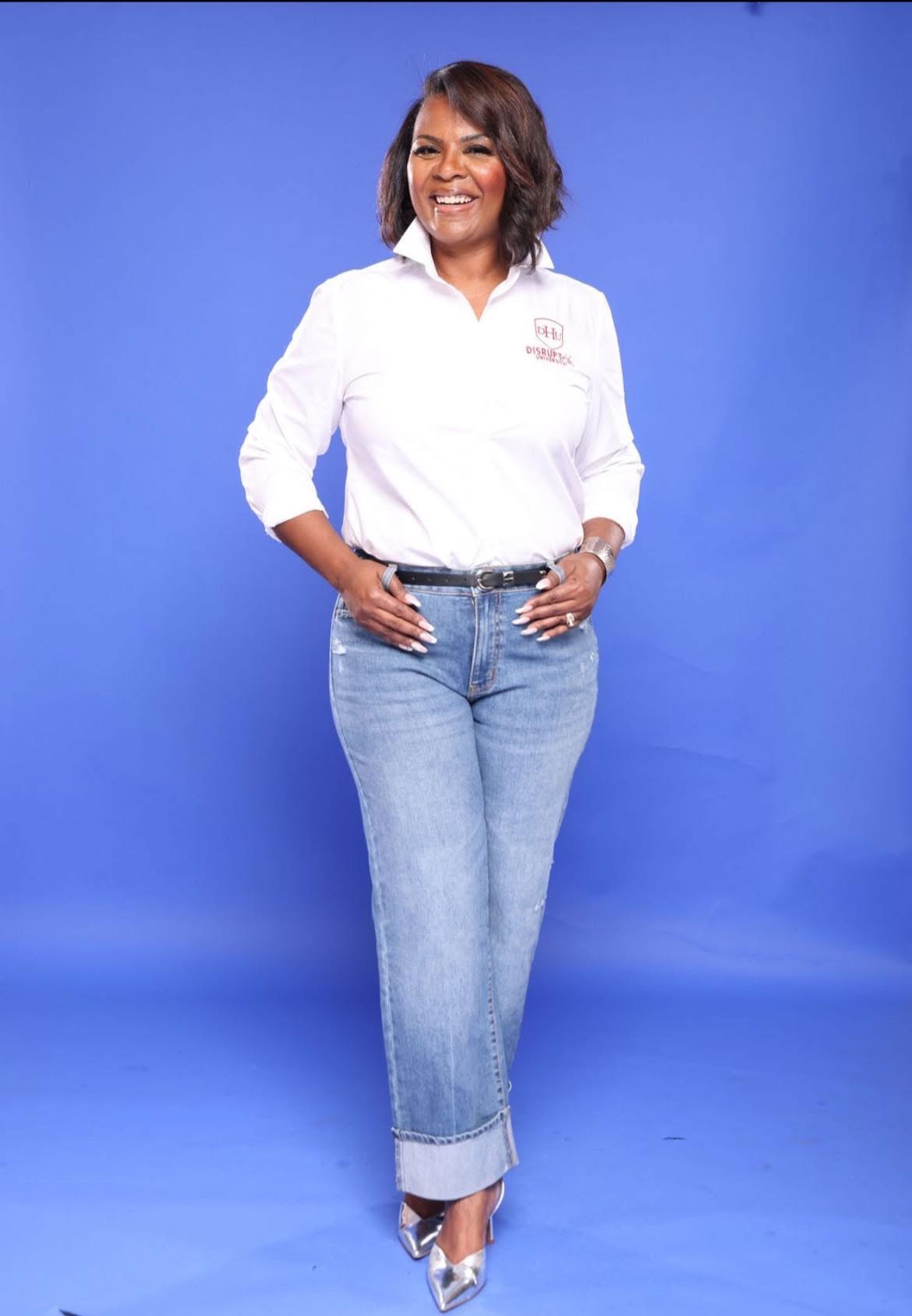 Woman standing with hands on hips, smiling, wearing white shirt and jeans, against blue background.