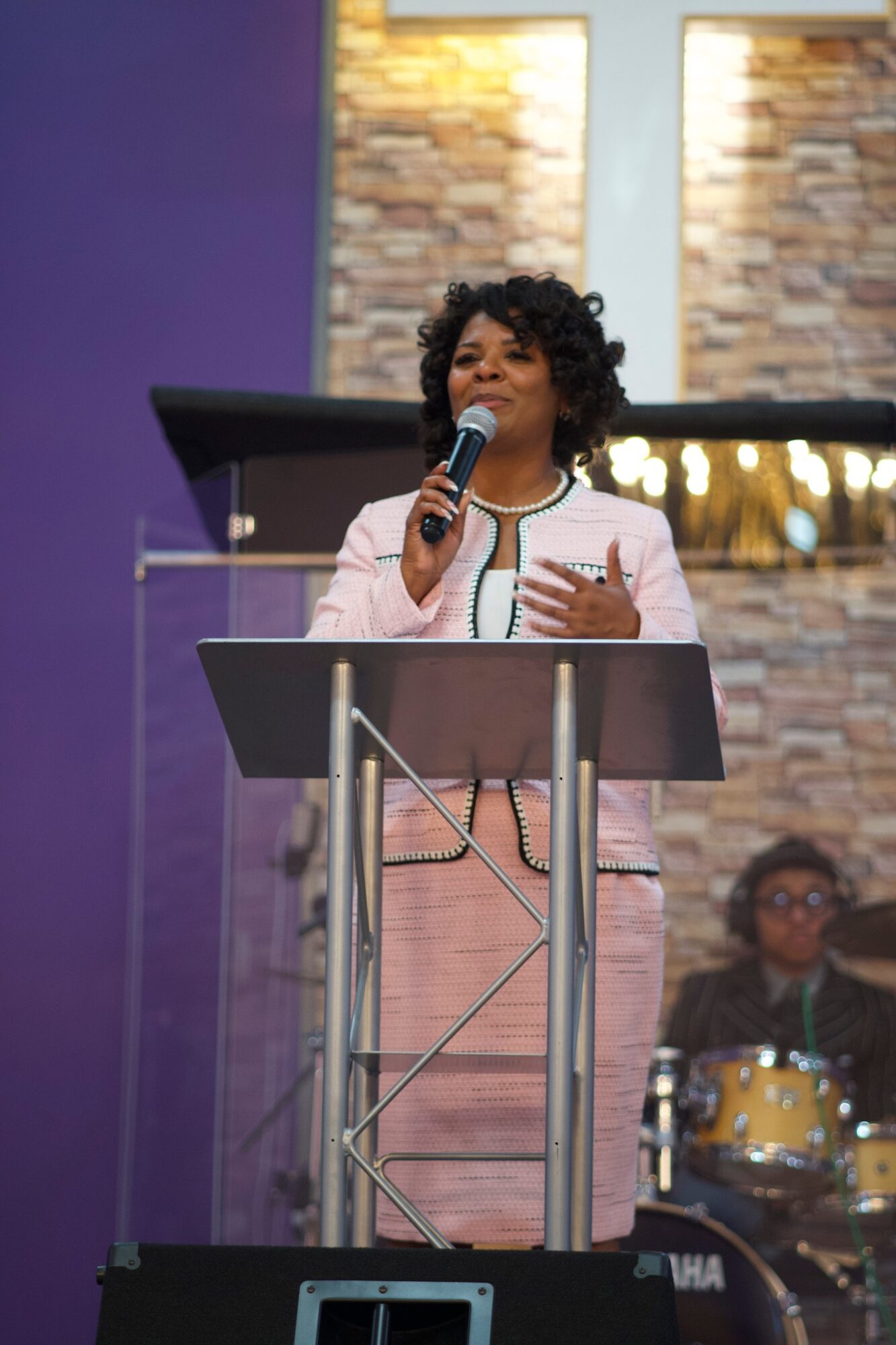 Woman with curly hair speaking into microphone at podium, background with brick wall and purple wall, musician playing drums in background.