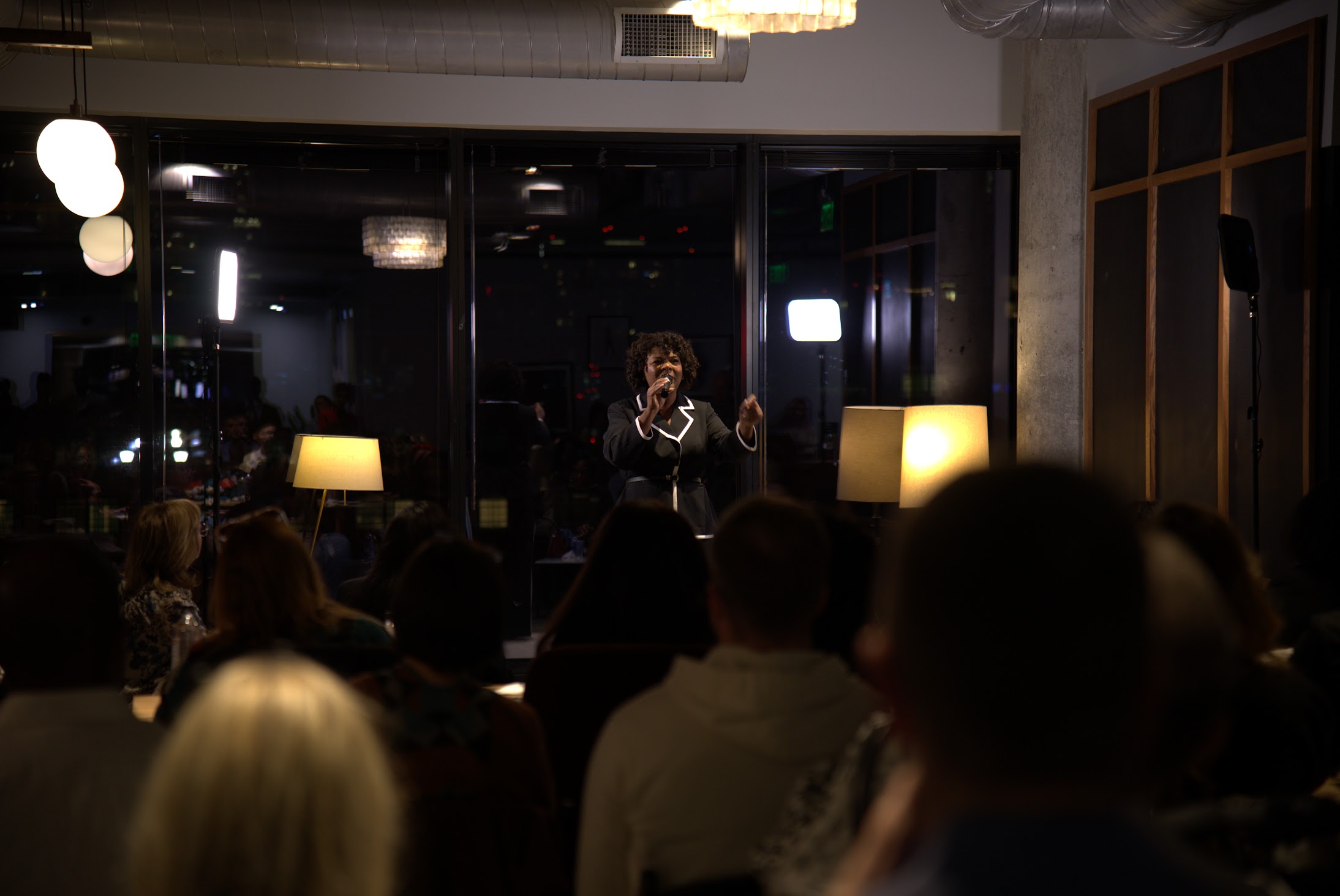 Person on stage speaking into microphone in front of seated audience in dimly lit room.