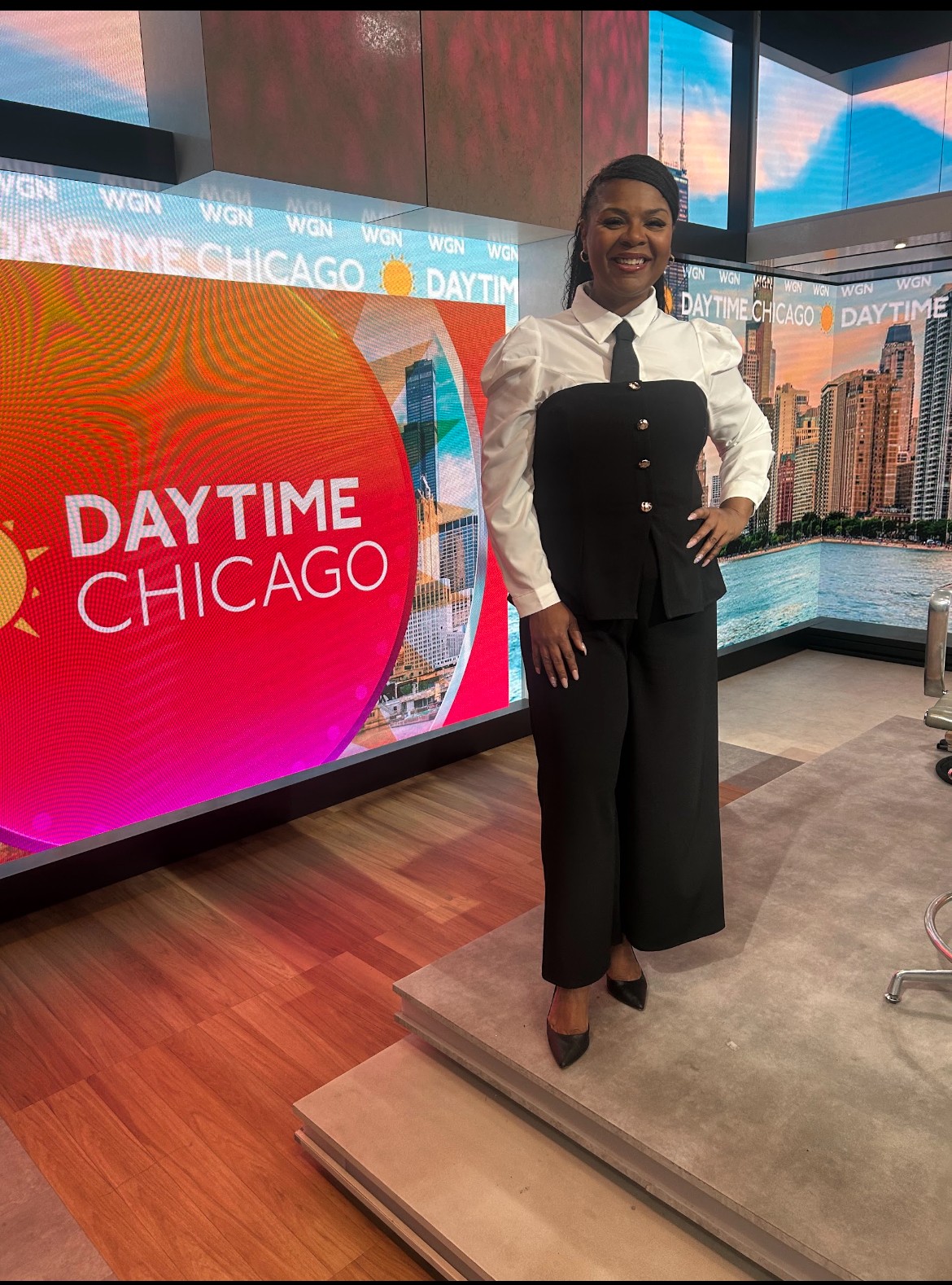Woman in black pants and white shirt standing on a stage with a cityscape background and a large screen displaying 'DAYTIME CHICAGO'.
