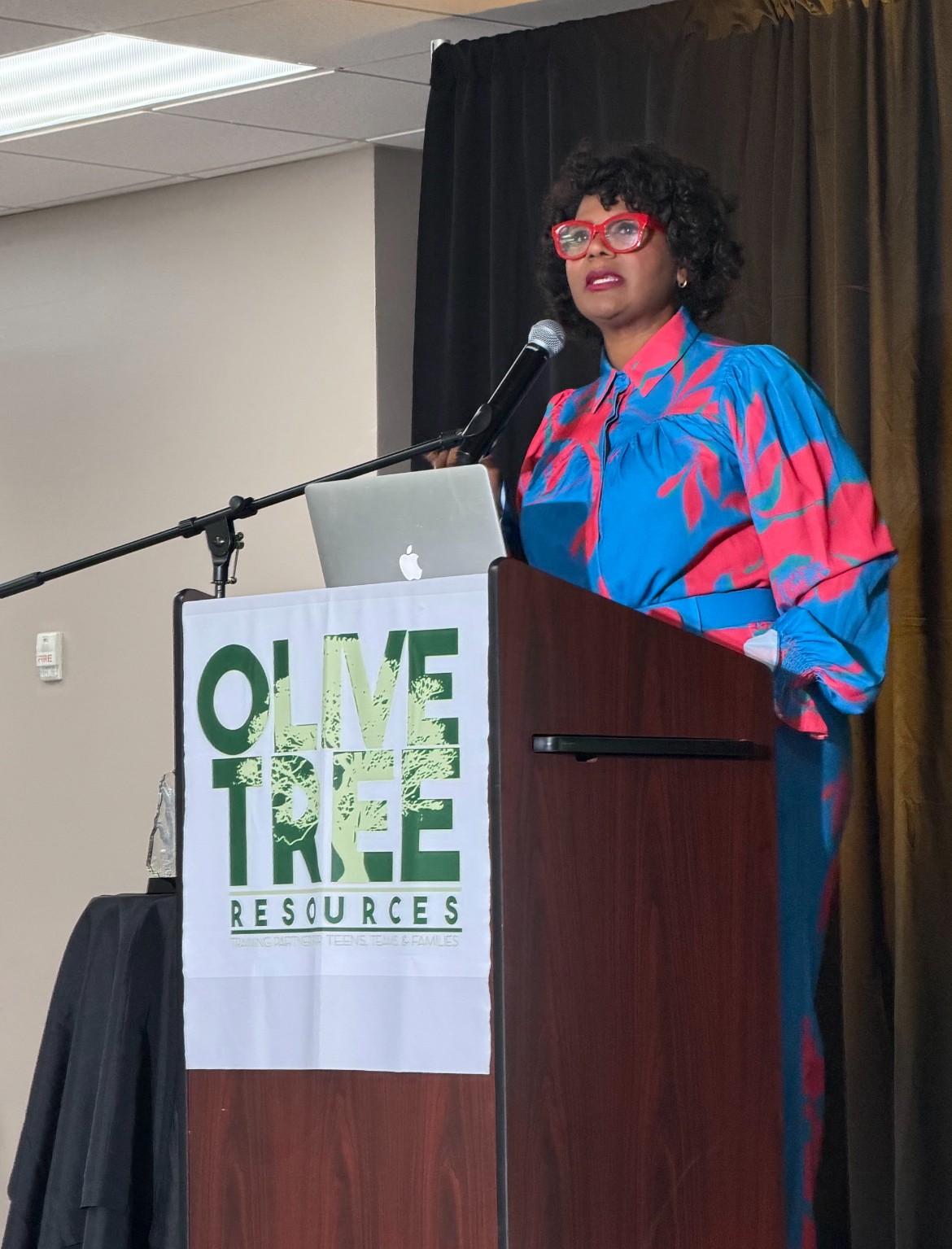 Woman with curly hair and red glasses speaking at a podium with a laptop, in front of a black curtain.