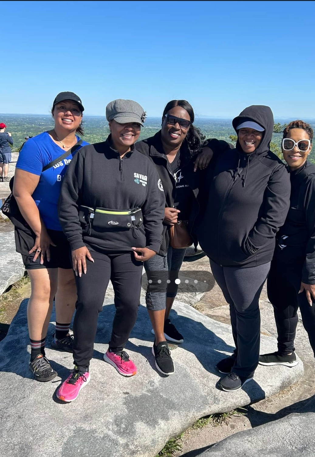 Group of six women standing outdoors on a sunny day, smiling, with a blue sky background.