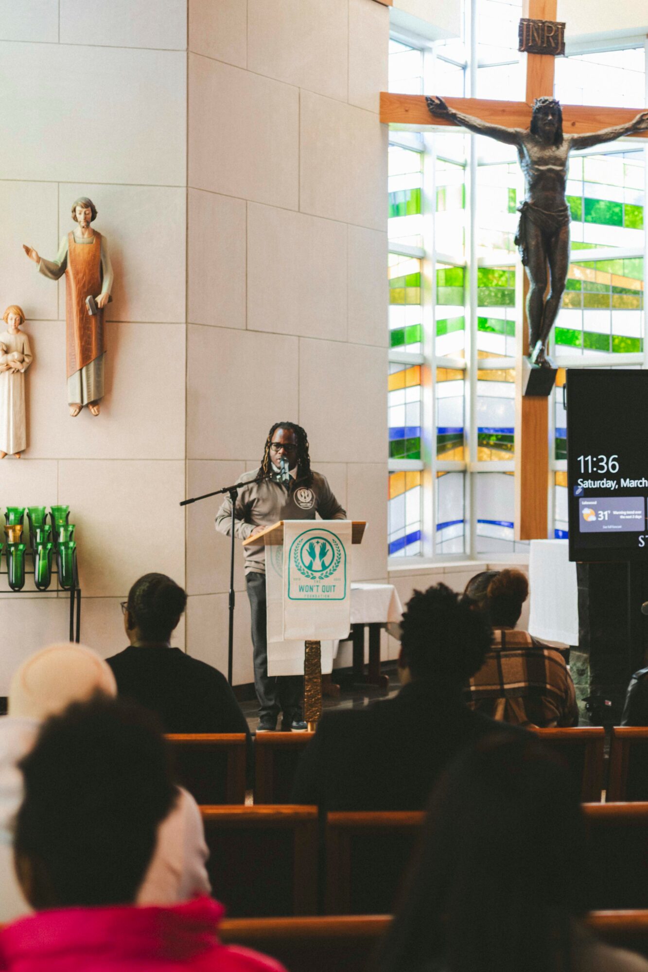 Person speaking at podium in church with crucifix and religious statues behind, audience seated in pews.