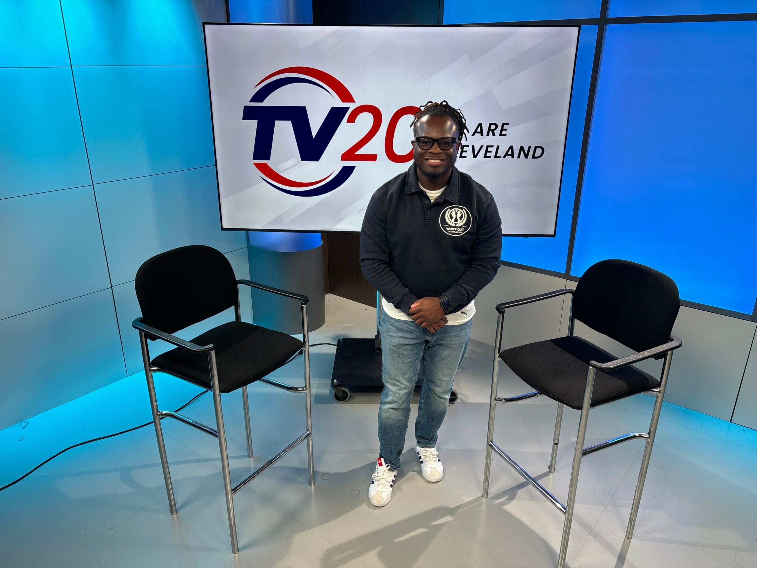 Person standing between two chairs in a studio with TV20 Cleveland logo on screen behind.