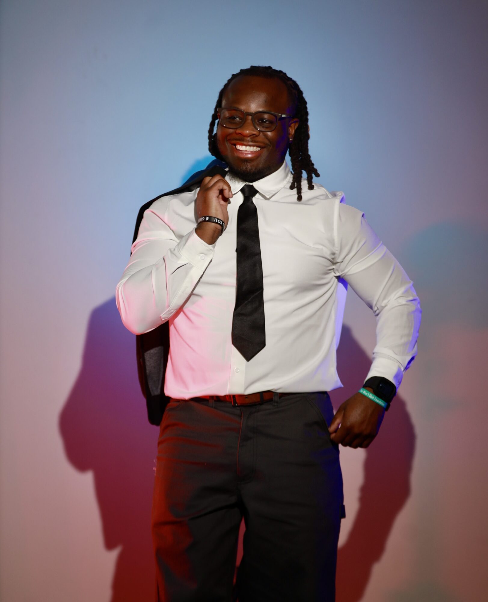 Person with glasses, dreadlocks, white shirt, black tie, black pants, smiling, holding a backpack strap, standing against a colorful background.
