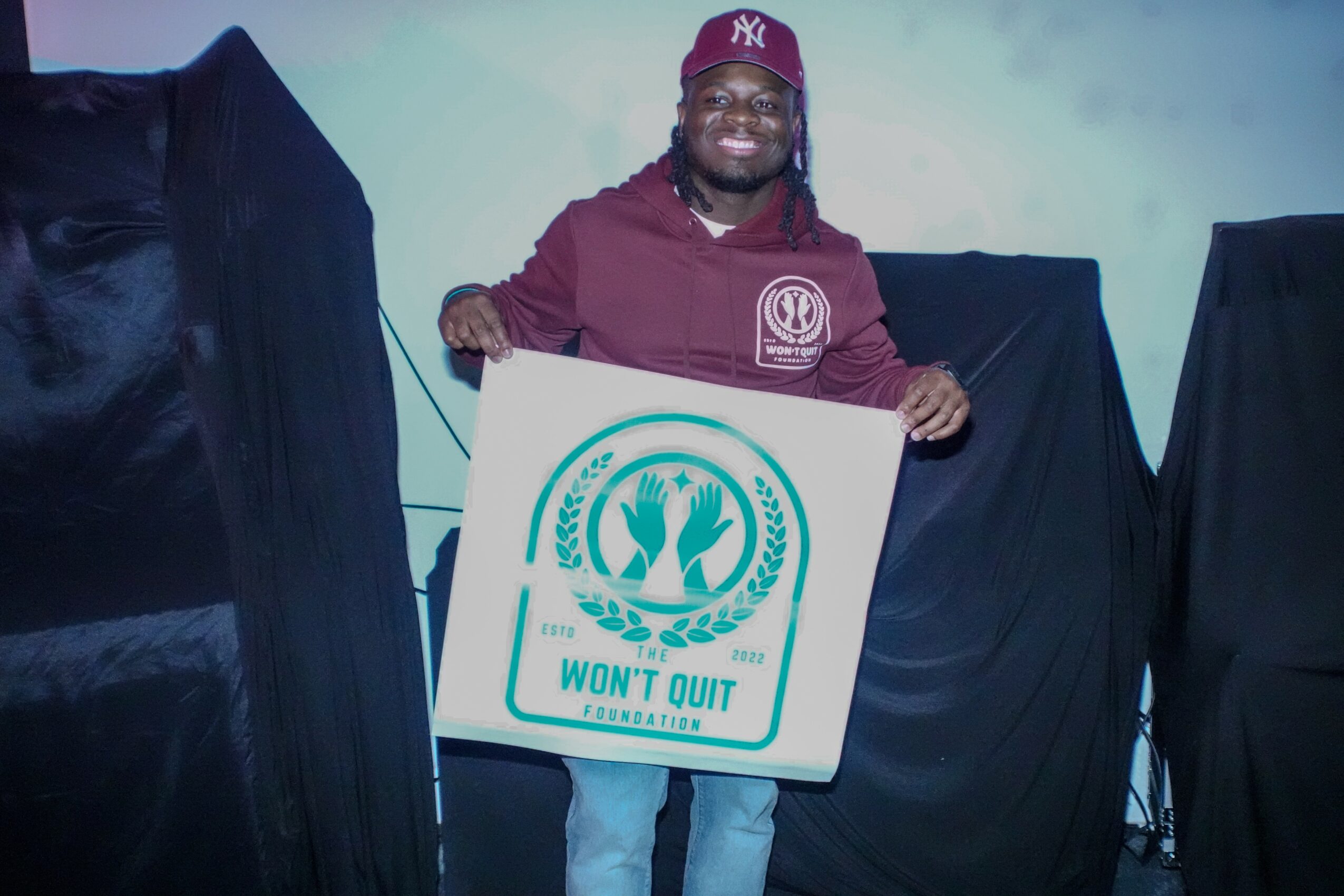 Person smiling holding a sign with a logo and text, standing between two black-covered objects.