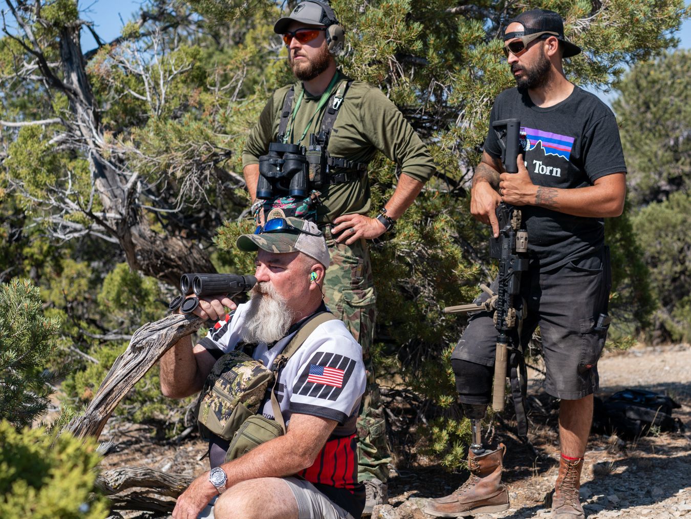 Three men outdoors among trees, one sitting and aiming a rifle, two standing with rifles, wearing casual and tactical clothing.