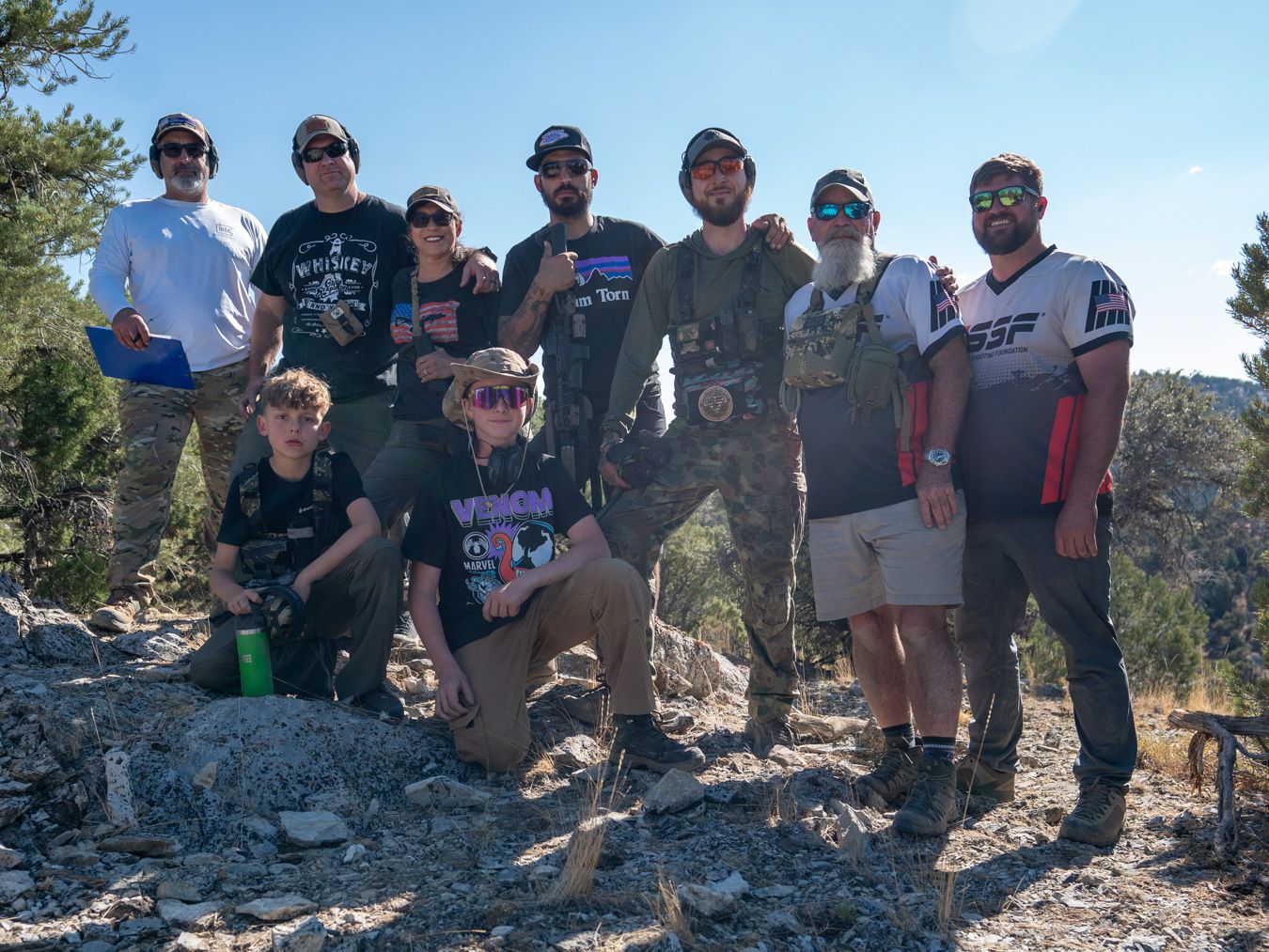 Group of ten people outdoors on rocky terrain with trees and blue sky in background.