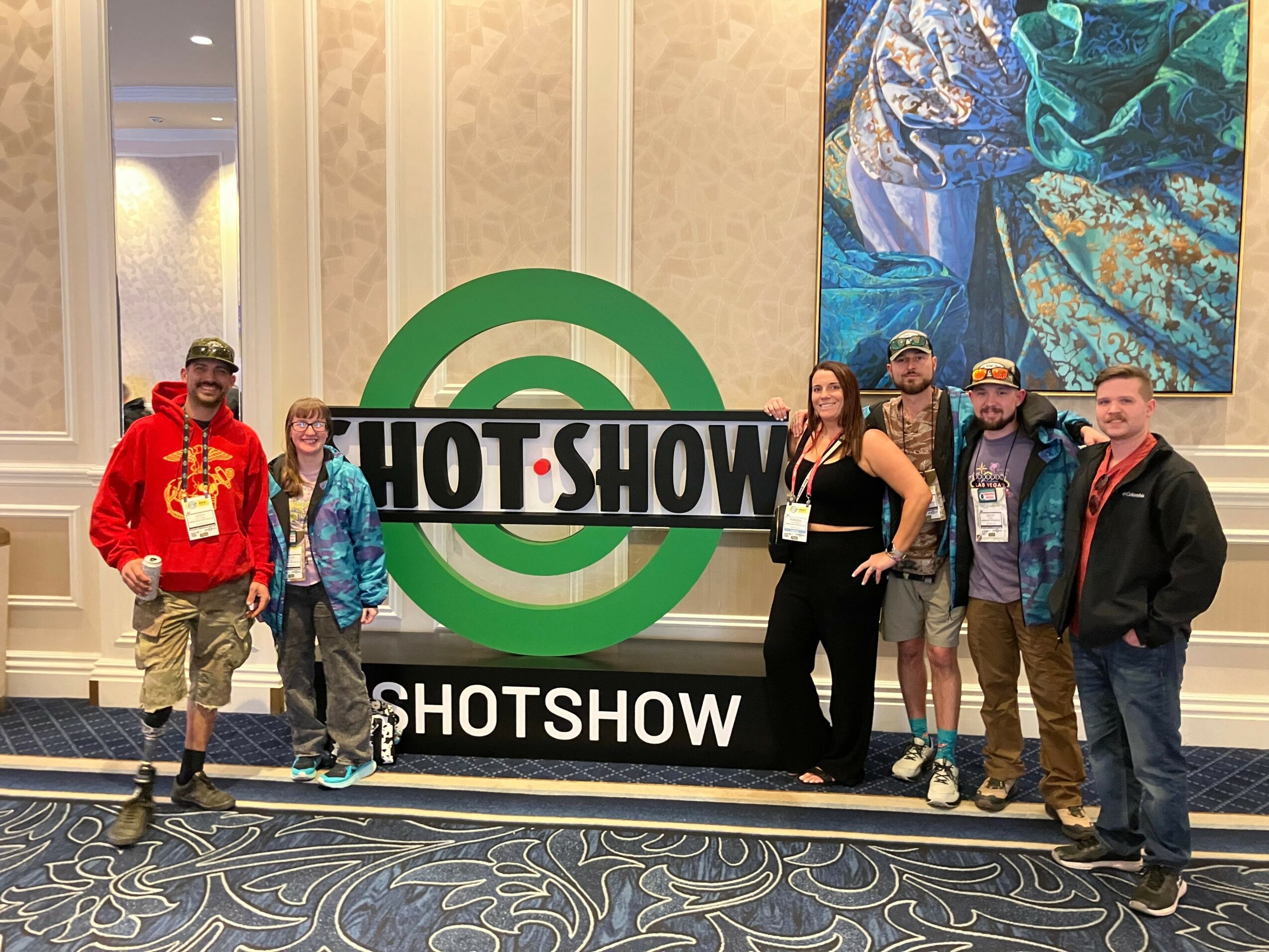 Group of six people standing in front of a large HOT SHOW sign indoors, with patterned carpet and artwork on wall.