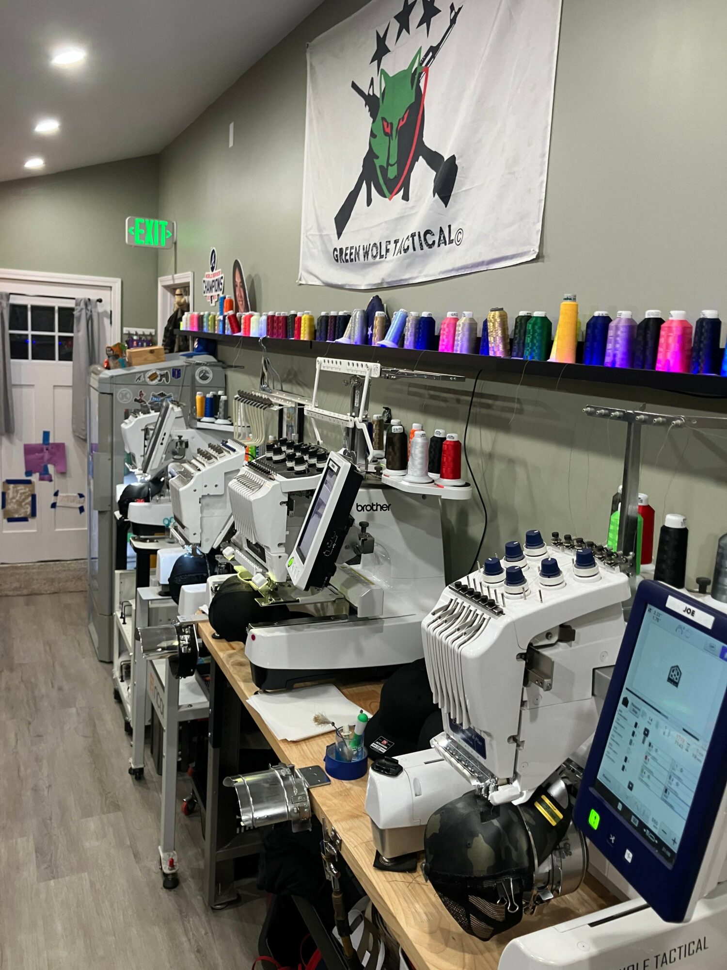 Sewing machines and spools of thread on shelves in a workspace with a logo banner on the wall.