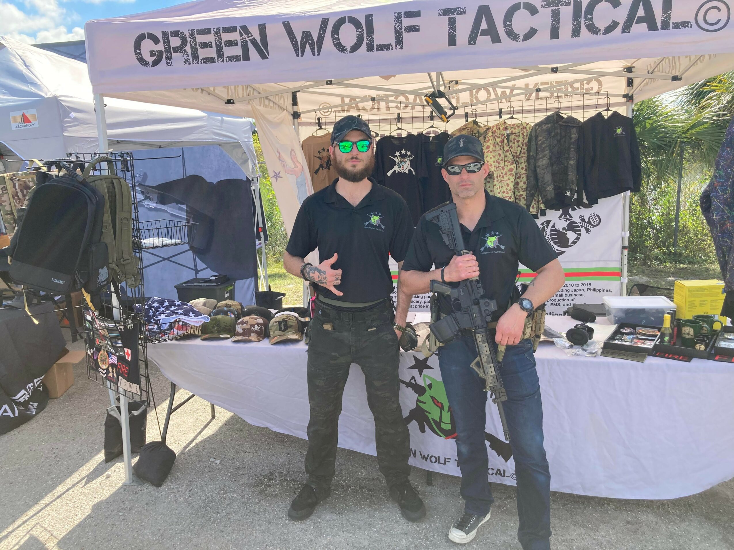 Two men stand in front of a tent with tactical gear and accessories displayed on tables and racks.