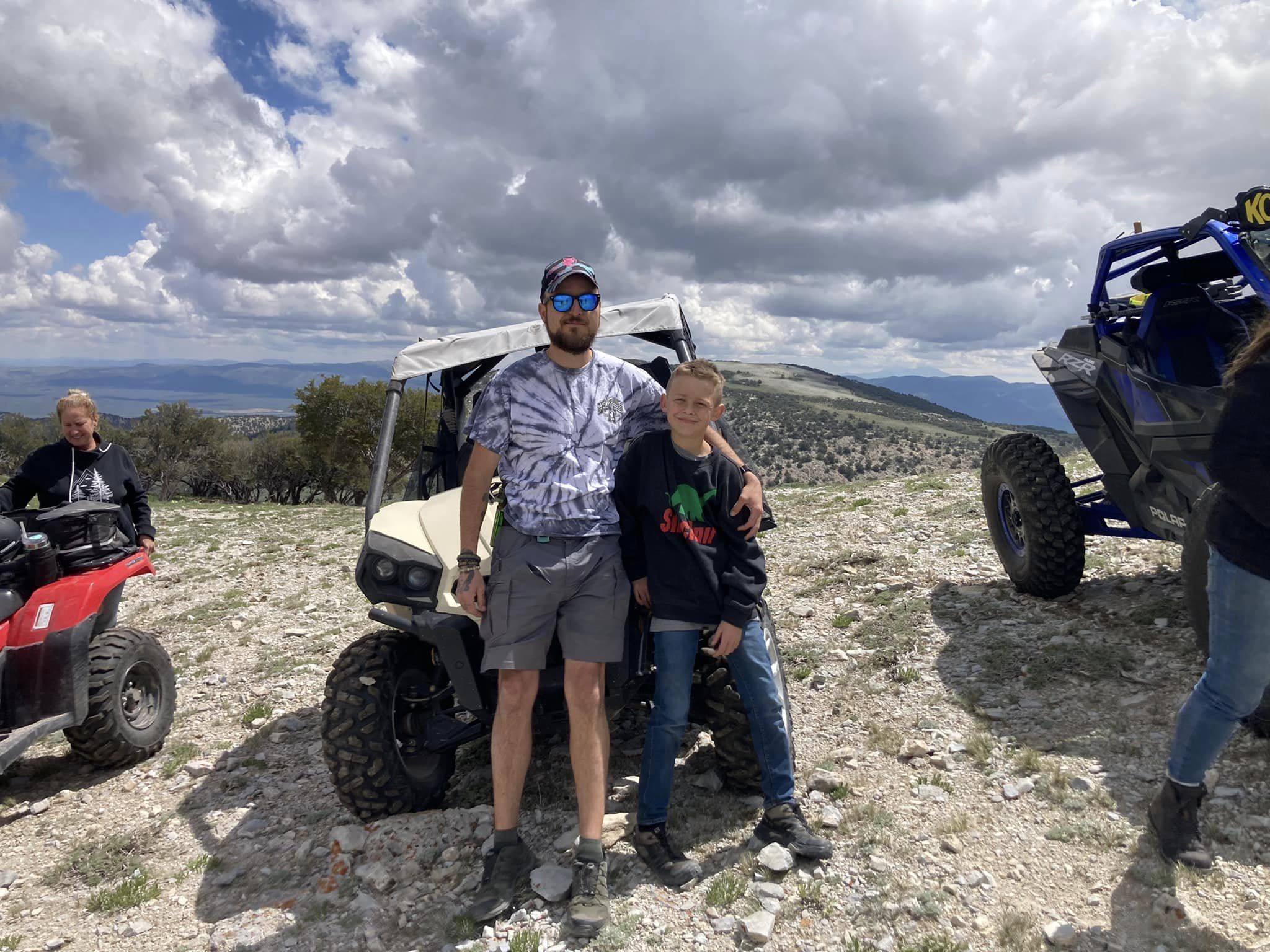 Two people standing outdoors on rocky terrain with a golf cart and off-road vehicles, cloudy sky in background.