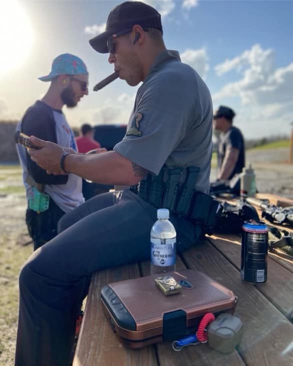Police officer sitting outdoors, looking at a phone, with a water bottle and equipment on a table nearby.