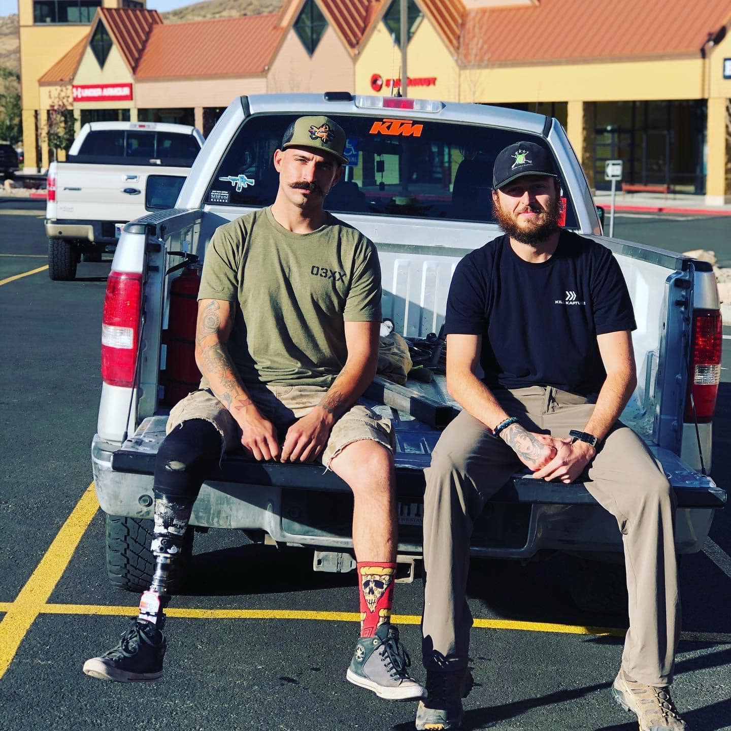 Two men sitting on the back of a pickup truck in a parking lot with stores in the background.