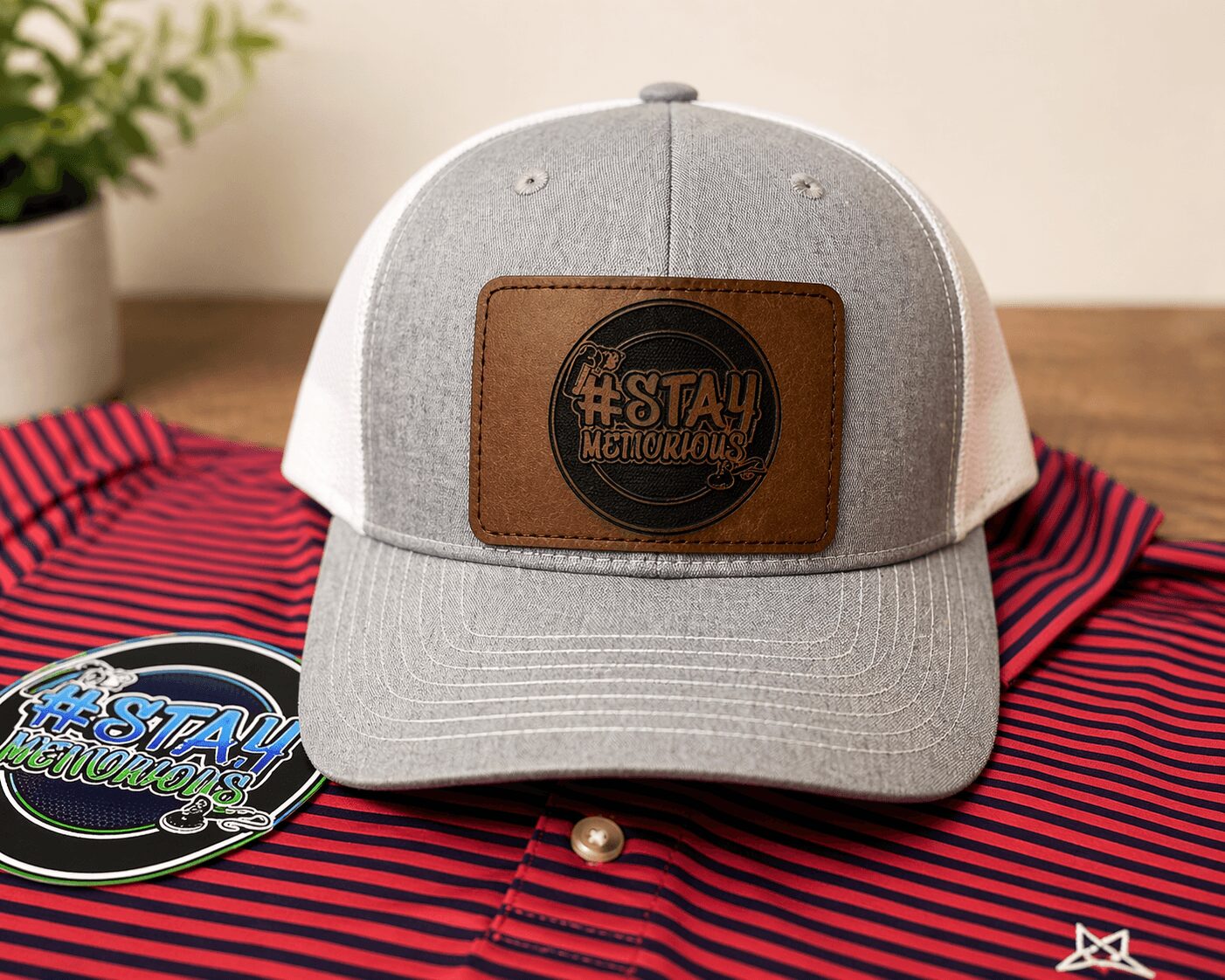 Gray and white trucker hat with brown patch on front, placed on red and black striped fabric, with a sticker nearby.
