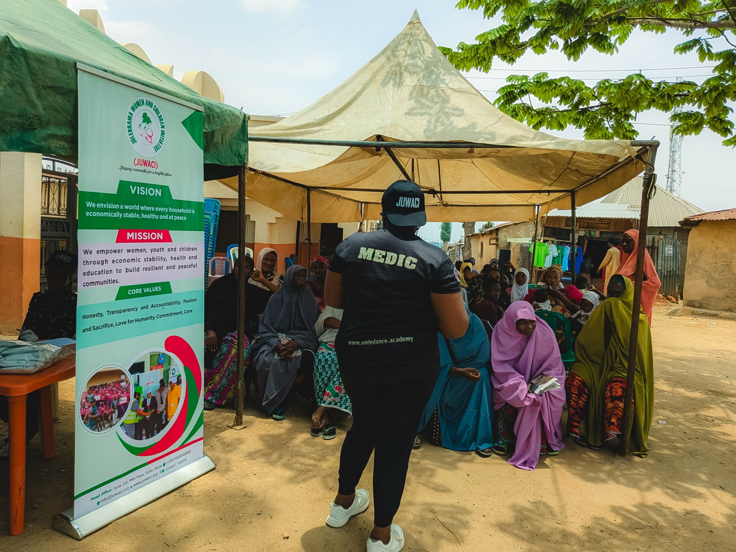 Medics and people gathered under a tent outdoors, with a banner on the left and trees in the background.