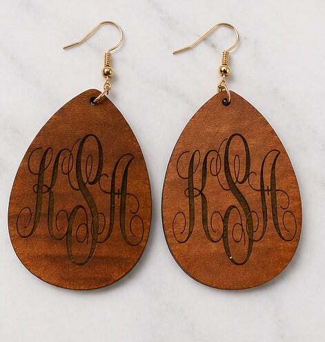 Pair of wooden earrings with engraved cursive initials, hooks attached at top, on a white background.