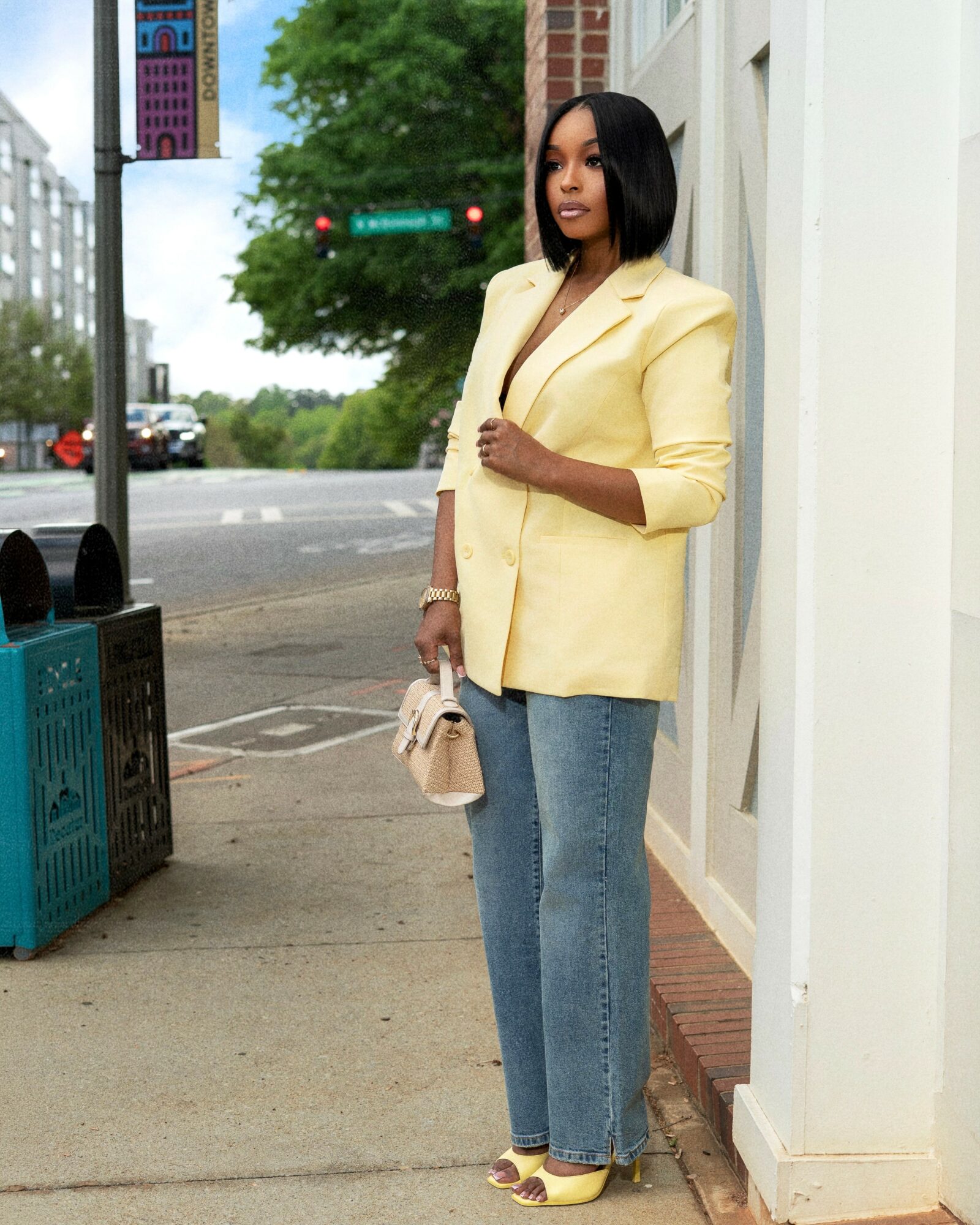 Woman in yellow blazer and jeans standing on sidewalk near white wall, holding purse, city street background.