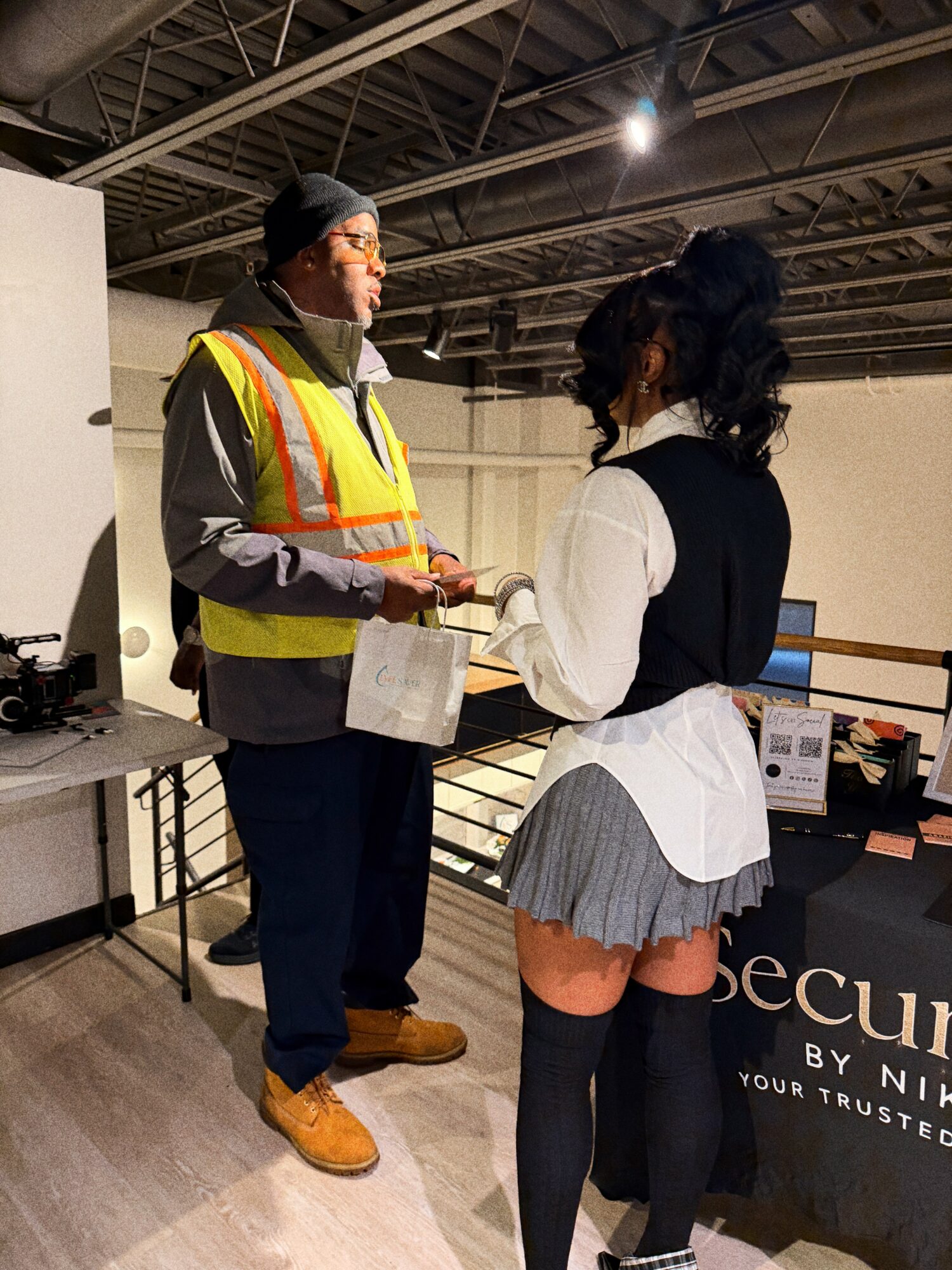 Two people talking indoors, one wearing a safety vest and the other in a white shirt and gray skirt.
