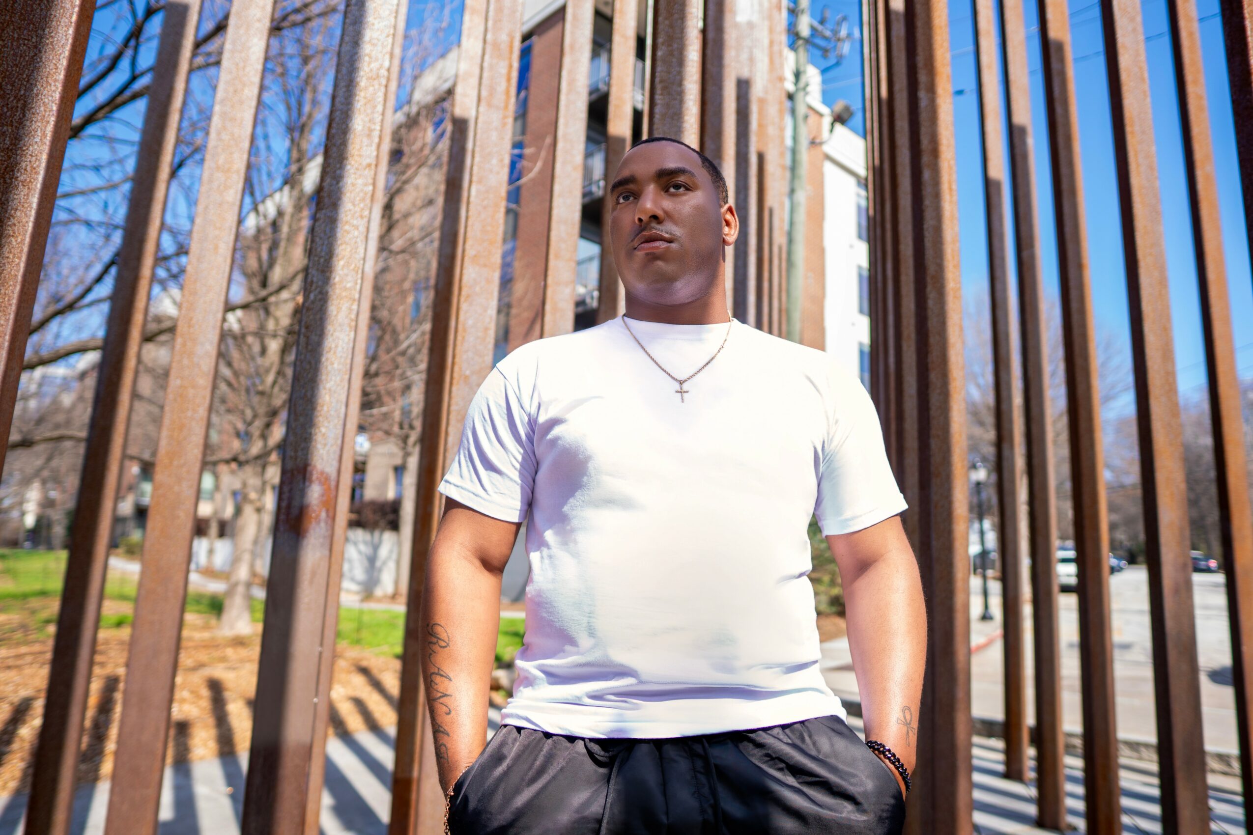 Man standing outdoors behind vertical metal bars, wearing a white t-shirt and dark pants, with trees and buildings in background.