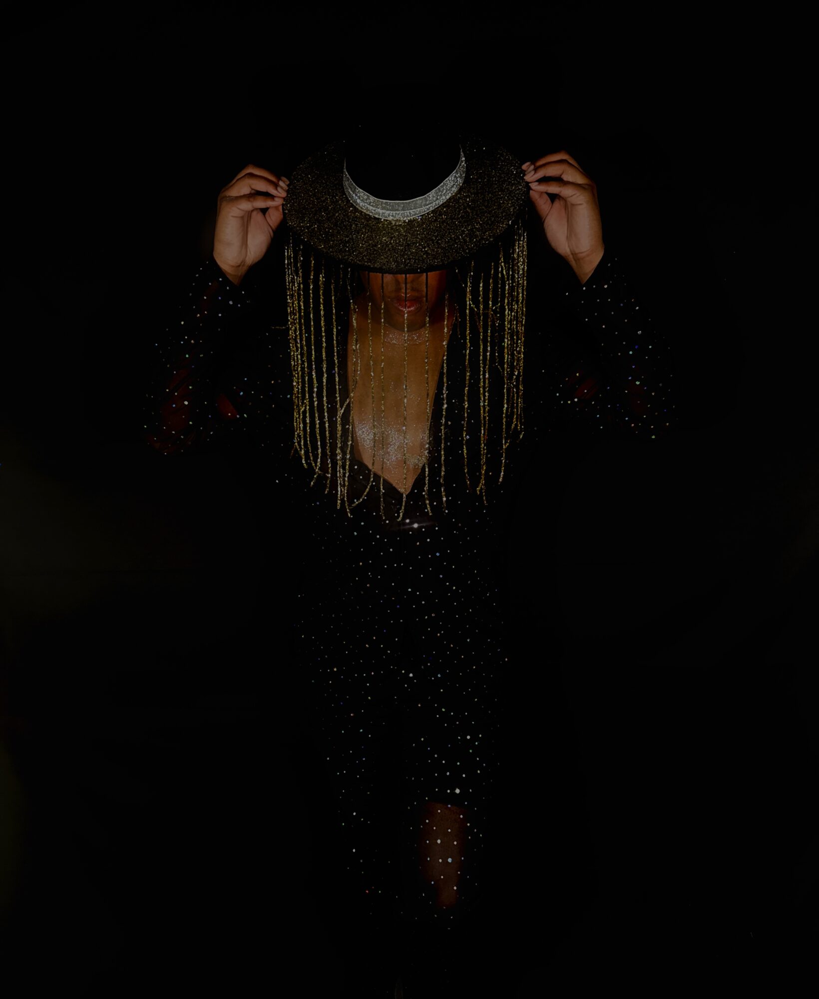 Person holding a hat with long gold chains, face obscured, dark background, wearing a dark outfit with small dots.
