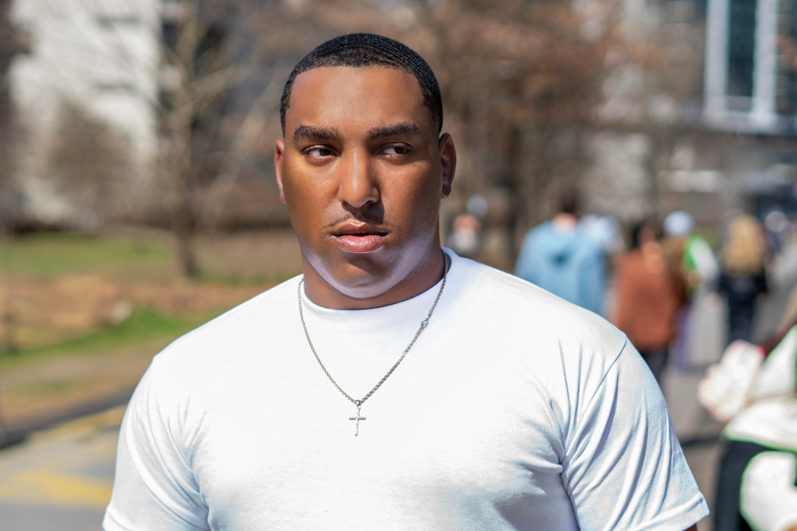 Young man with short hair wearing a white T-shirt and a cross necklace outdoors.