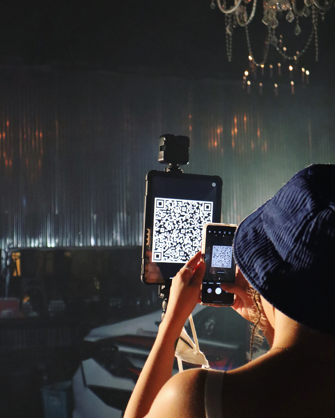 Person taking a photo with a smartphone, holding a QR code on a tablet, in a dimly lit setting with a chandelier.