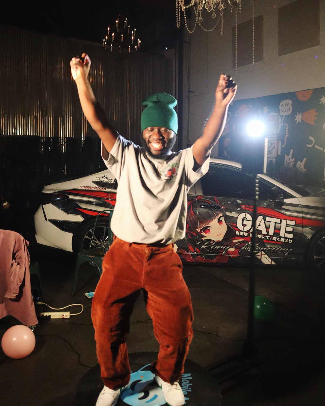 Person with arms raised, wearing a green beanie, gray shirt, and brown pants, smiling in a dimly lit room.