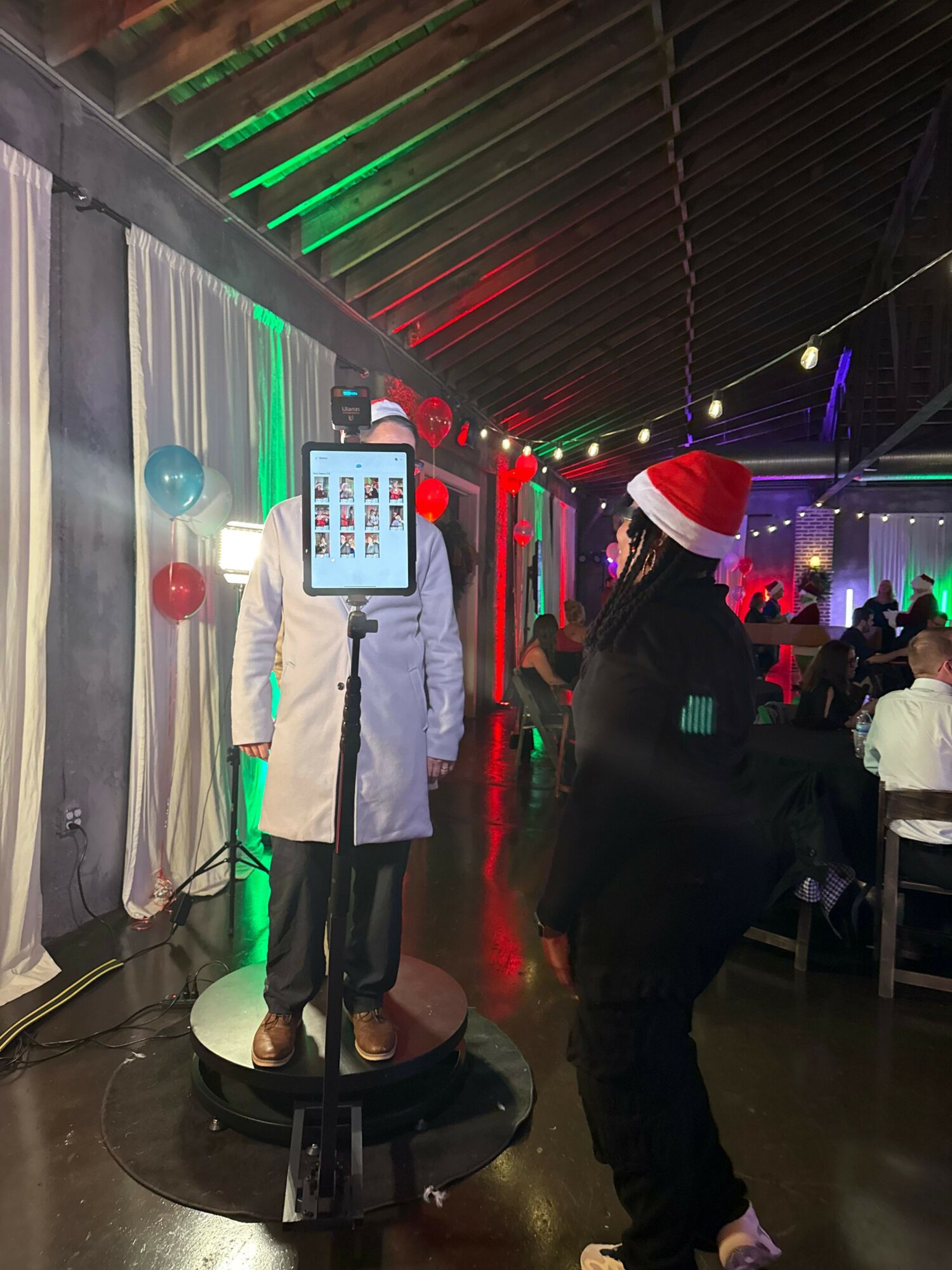 Person in white coat with a Santa hat standing near a device on a stand at a festive event.