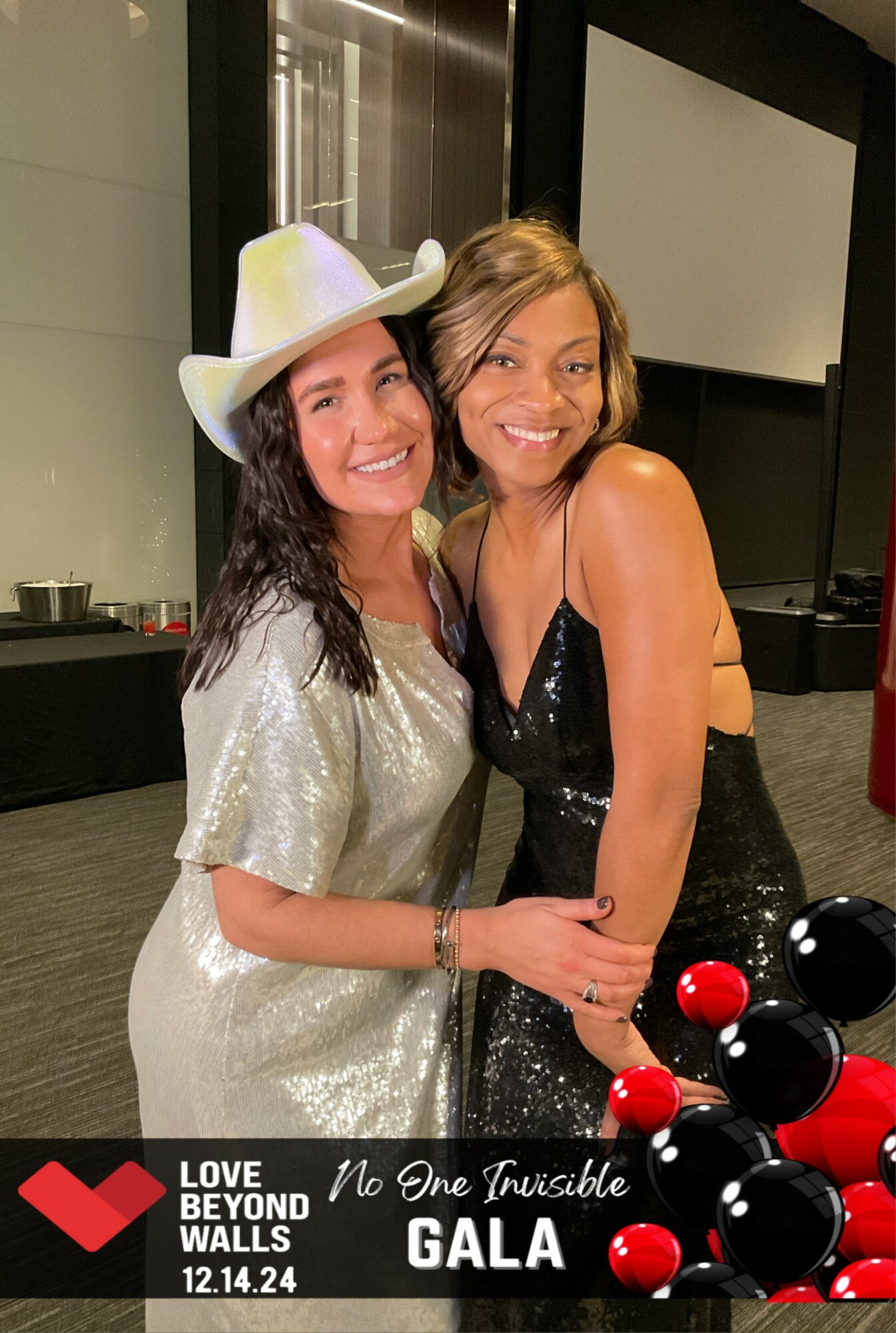 Two women smiling, one wearing a white hat and silver dress, the other in a black dress, holding hands.