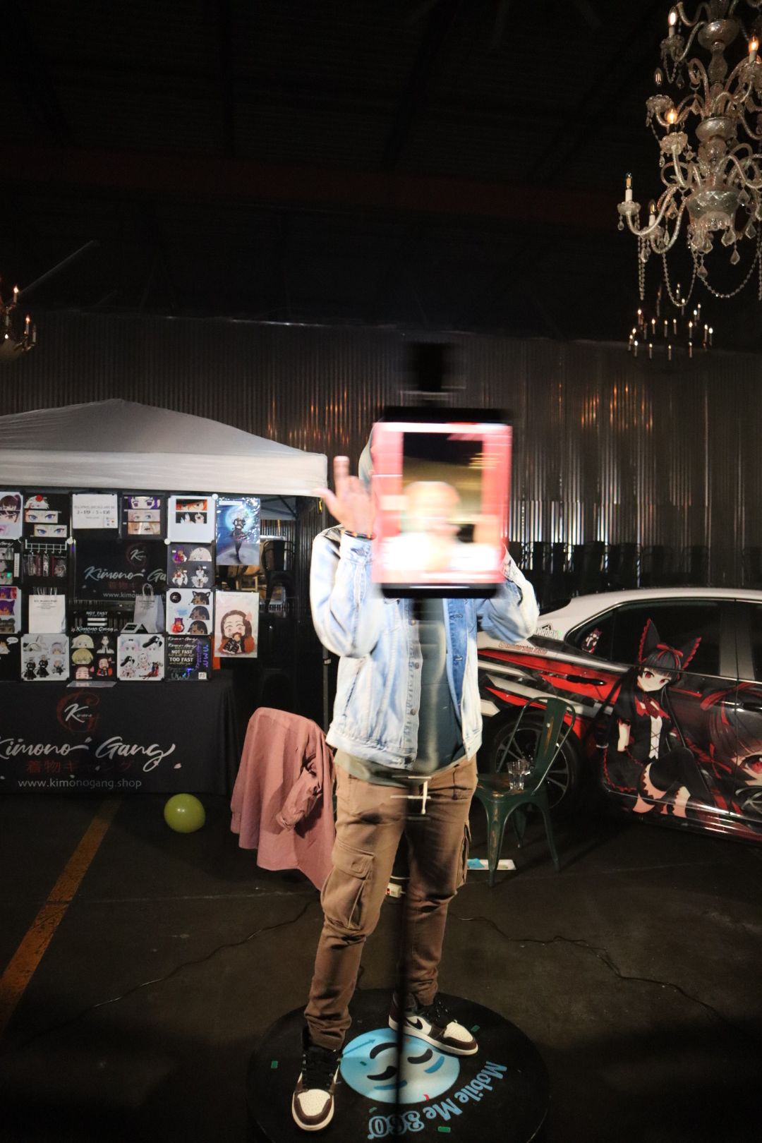 Person holding a tablet in front of face, standing indoors with chandeliers and a black curtain backdrop.