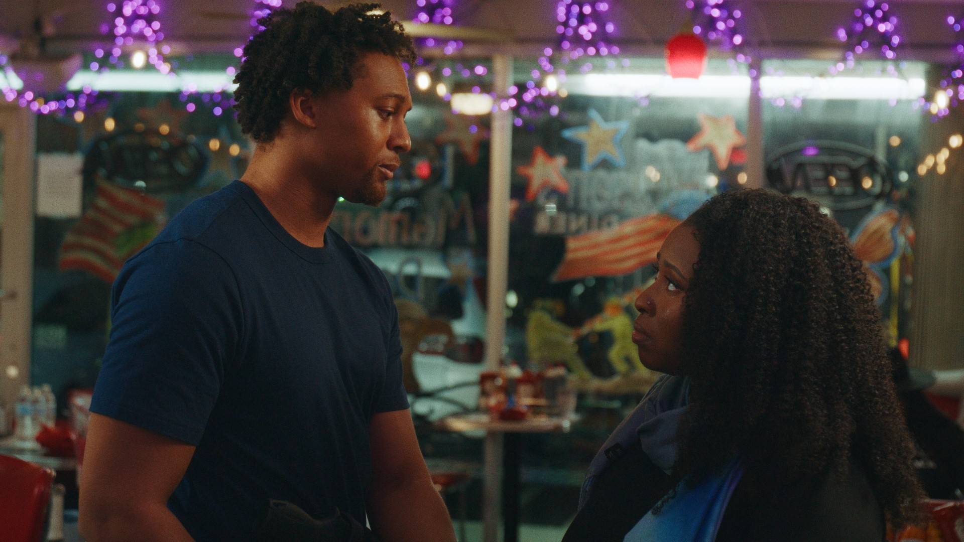 Two people facing each other in a colorful indoor setting with lights and decorations.