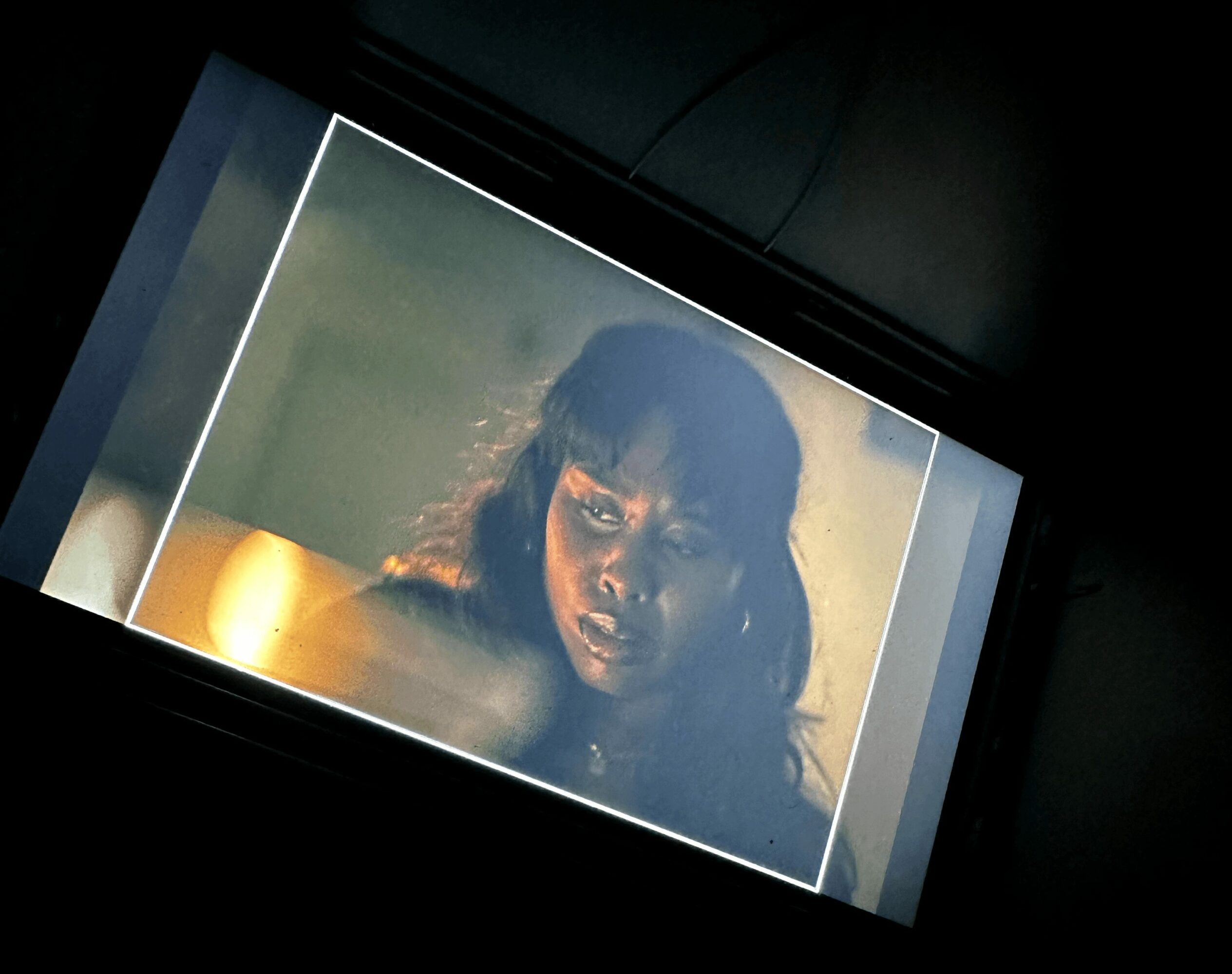 Screen displays a woman with long dark hair and intense expression, illuminated by a yellowish light, against a dark background.