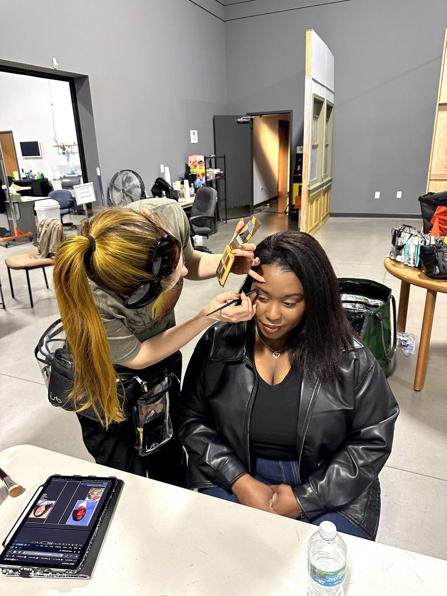 Makeup artist applying makeup to a seated woman in a room with chairs and tables.