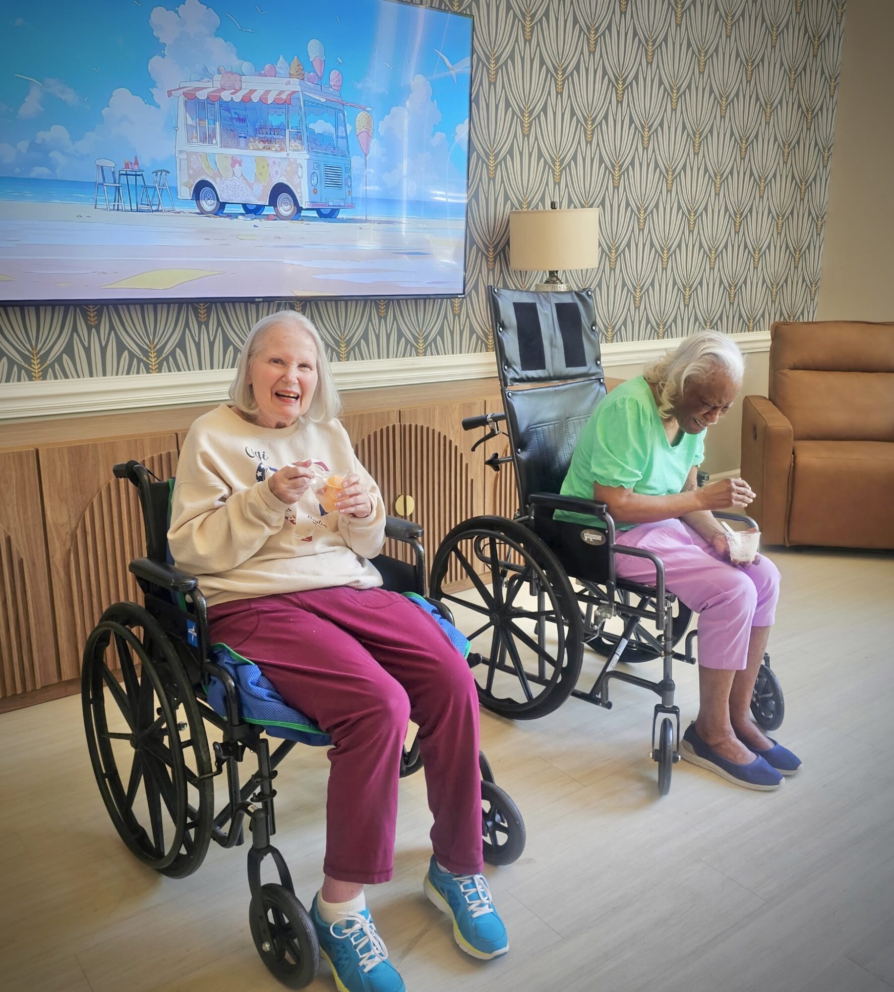Two elderly women in wheelchairs, one smiling and holding a cup, the other looking down at a cup, in a room with patterned wallpaper and a large TV screen.